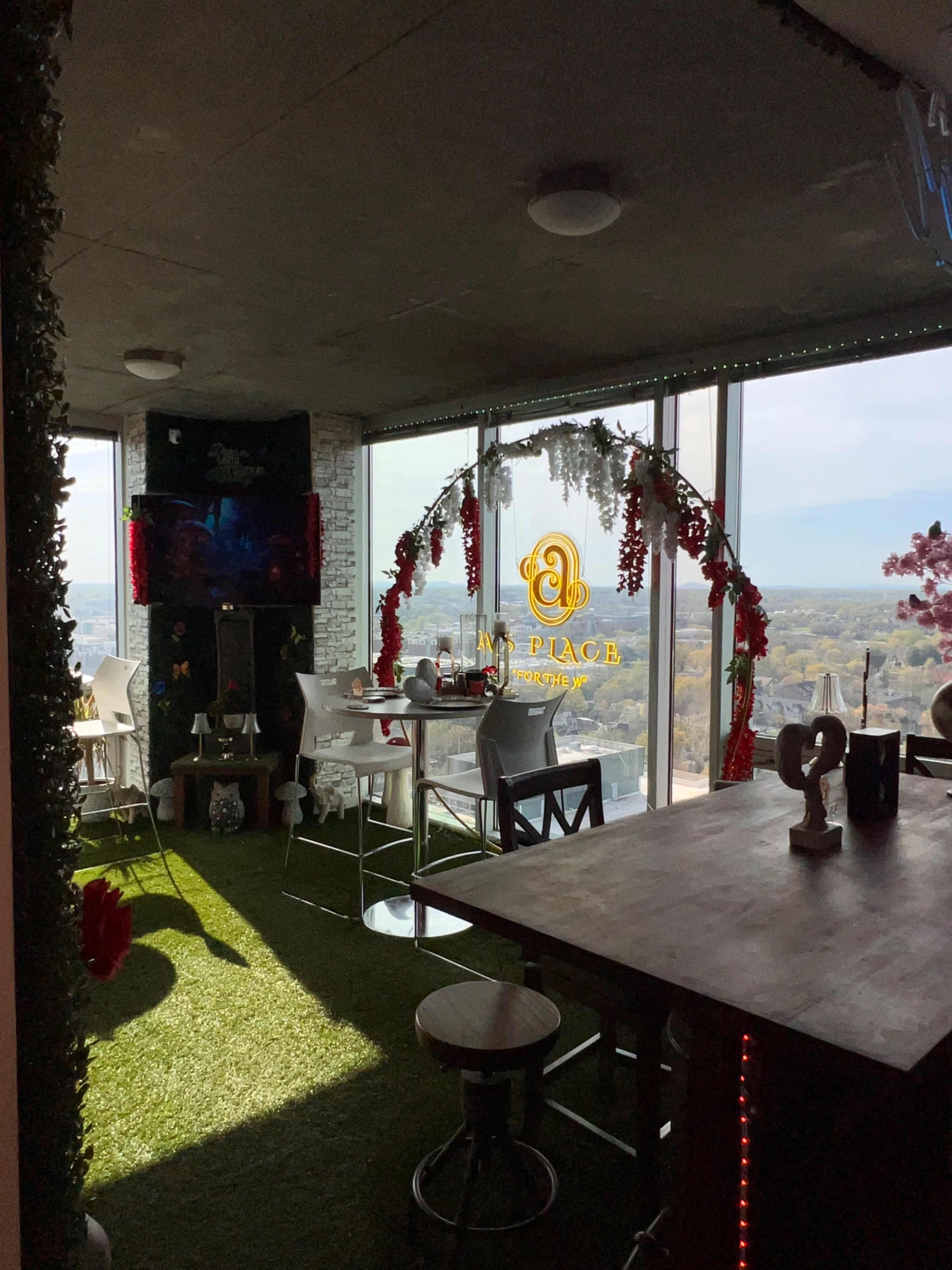 The image shows a rooftop restaurant with artificial grass, decorative flowers, and a large window offering a view of the outdoor scenery.
