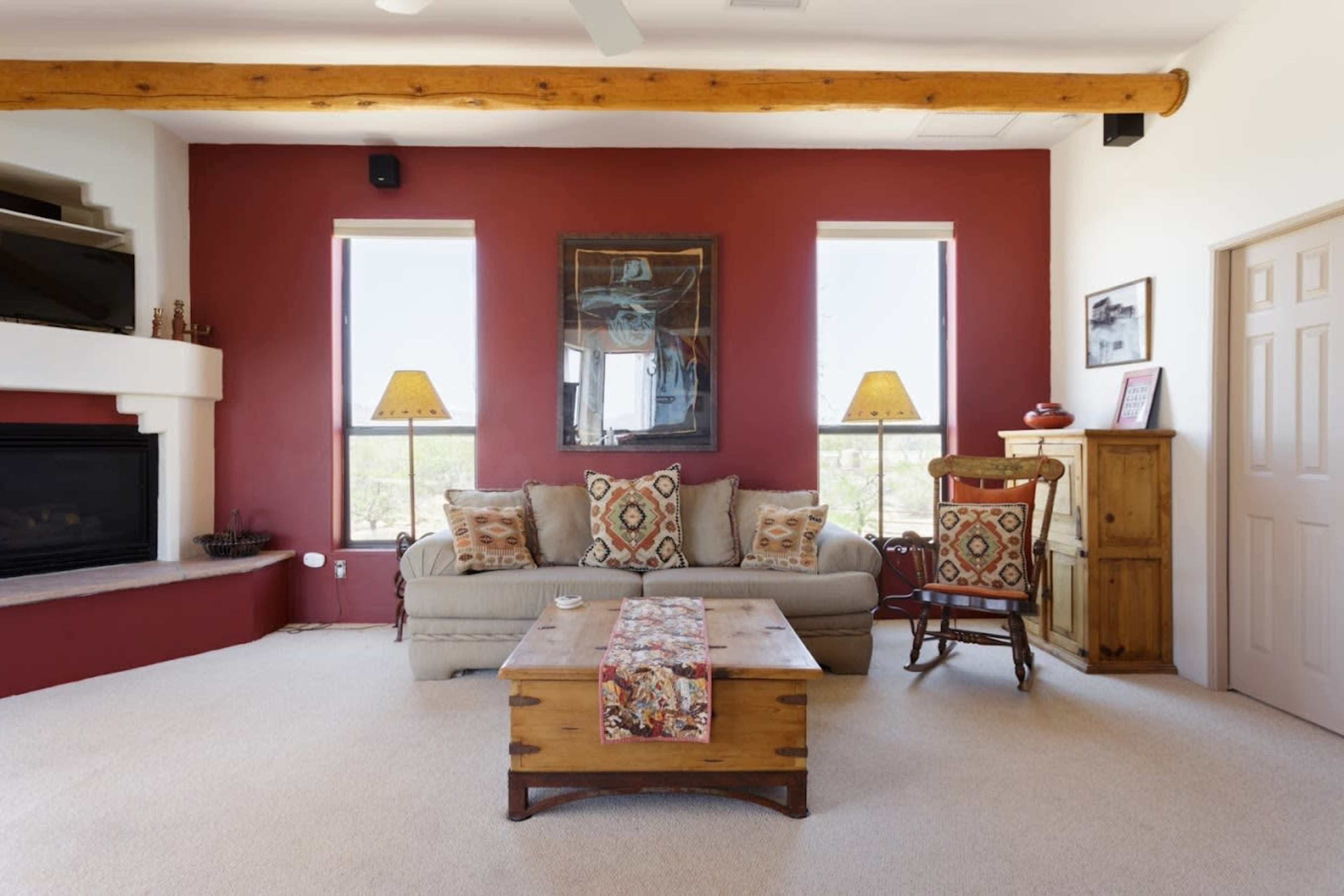A living room features a light-colored sofa with decorative pillows, a wooden coffee table, two lamps on window sills, and a rustic cabinet against a red accent wall.
