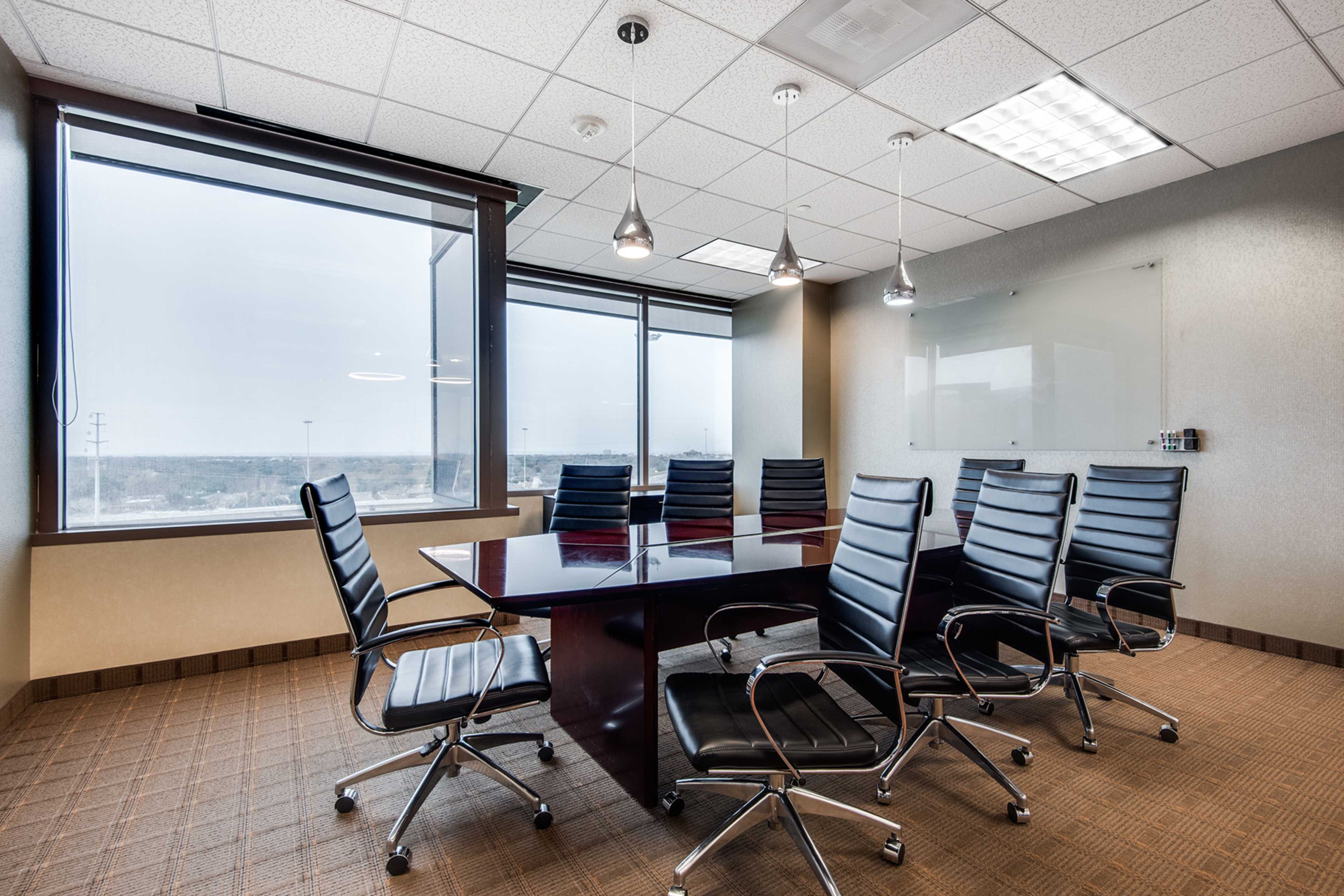 A modern conference room features a large rectangular table surrounded by black leather chairs, with large windows offering a view of the outside.