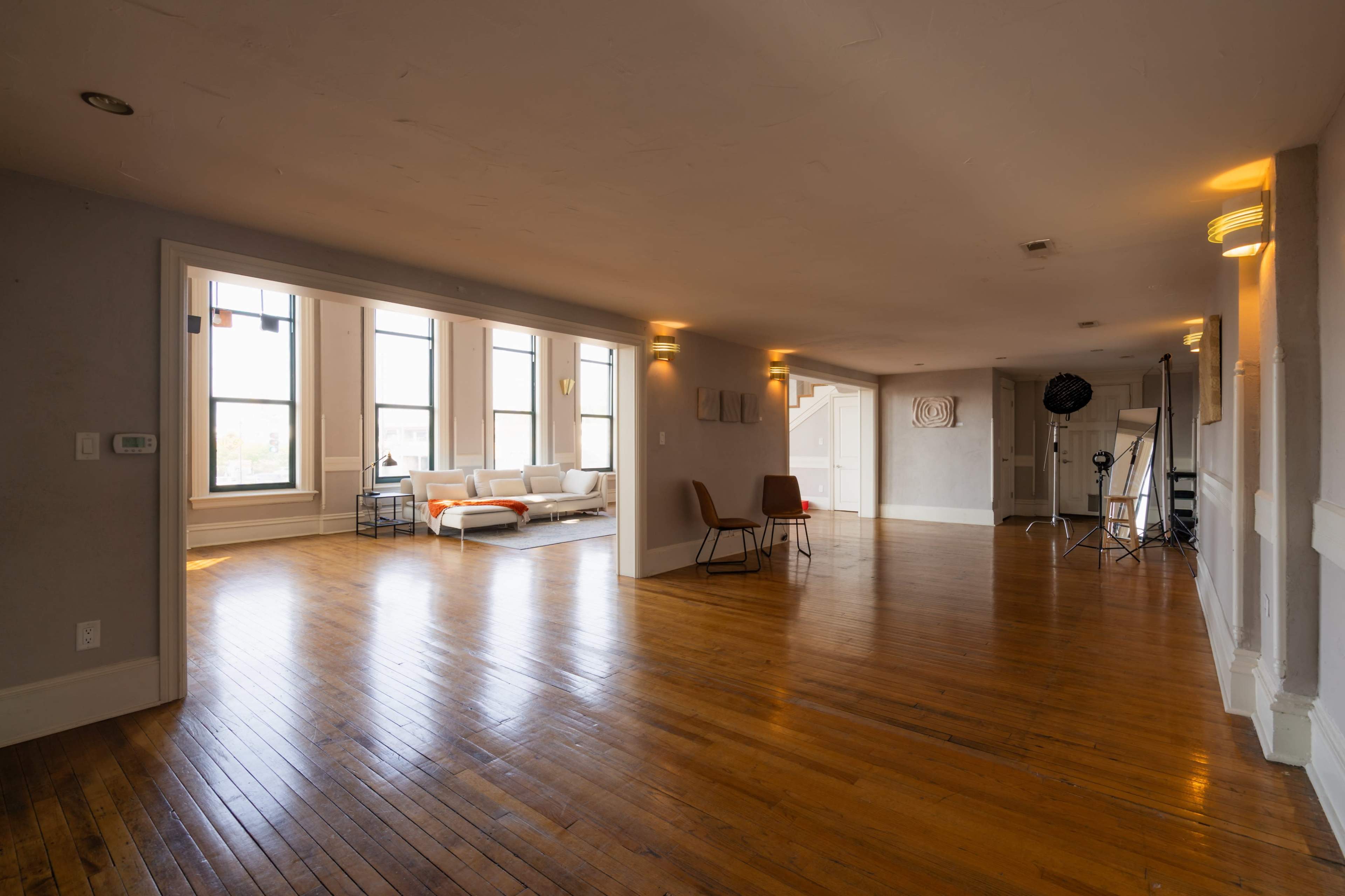 The image shows a spacious, well-lit room with hardwood flooring, featuring a seating area and large windows.
