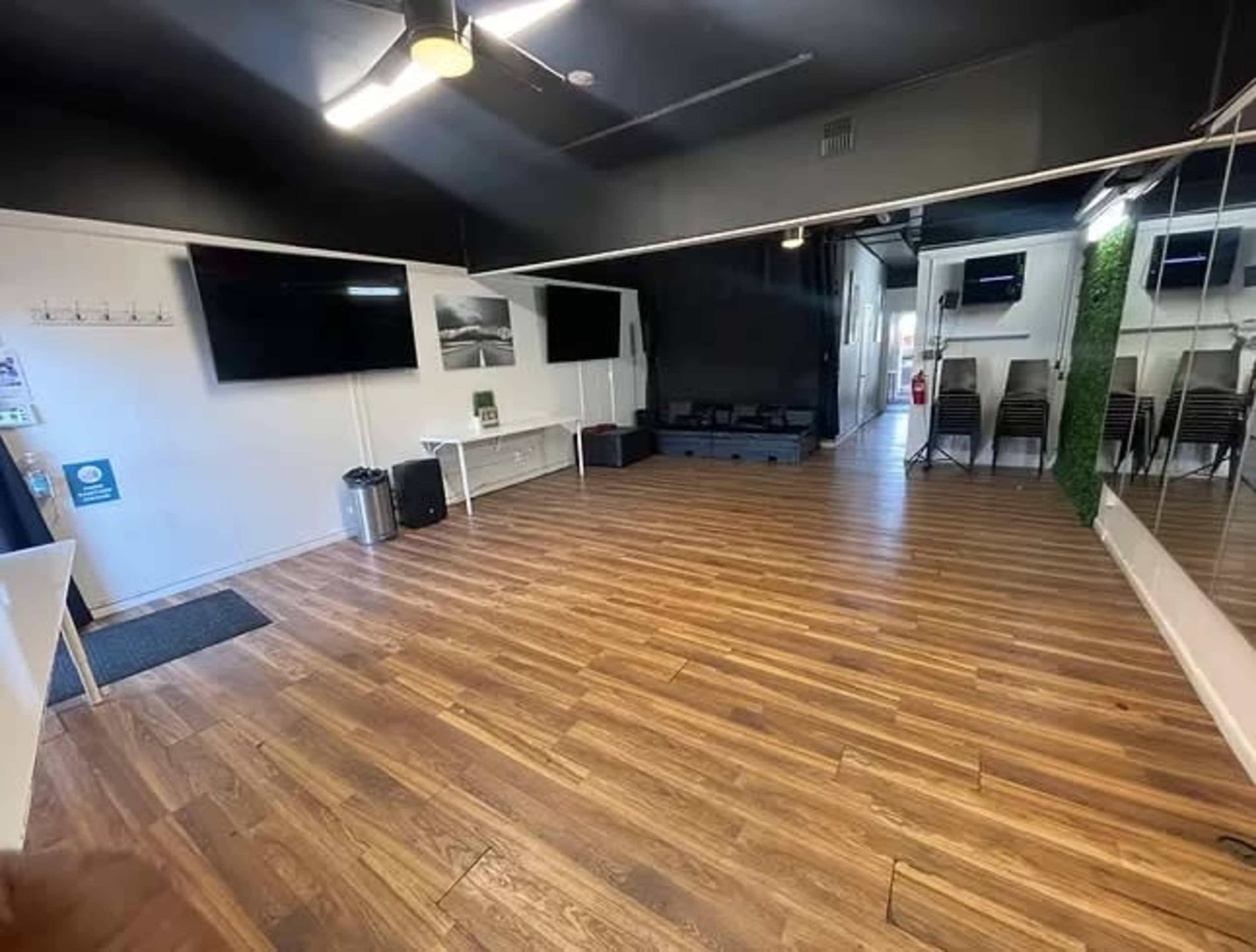 The image shows an empty room with wooden flooring, two large flat-screen TVs on the walls, and a mirrored section leading to a space with black seating and chairs.