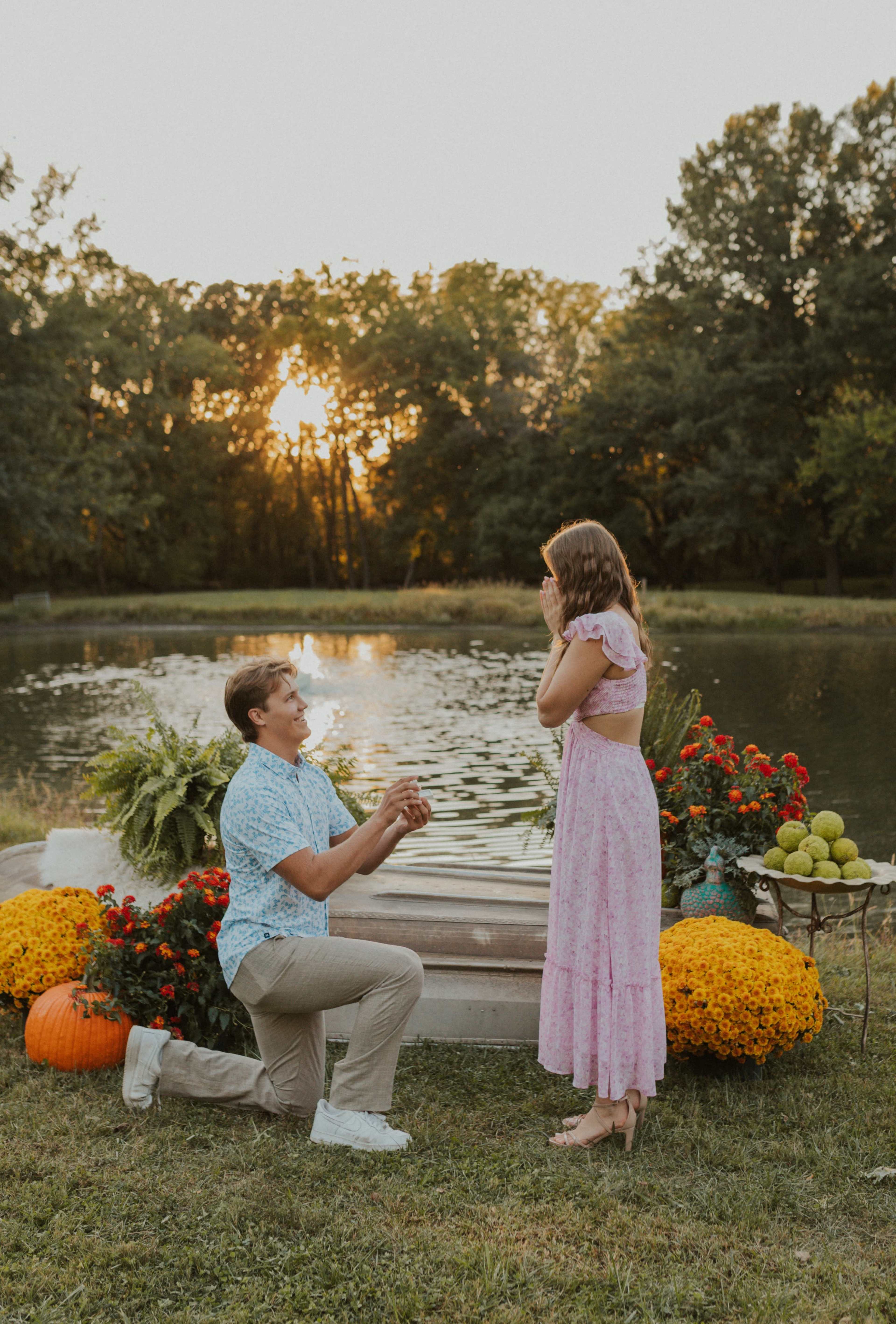 A man is kneeling with a ring in front of a woman, who is covering her face in surprise, by a pond surrounded by flowers and autumn decorations at sunset.
