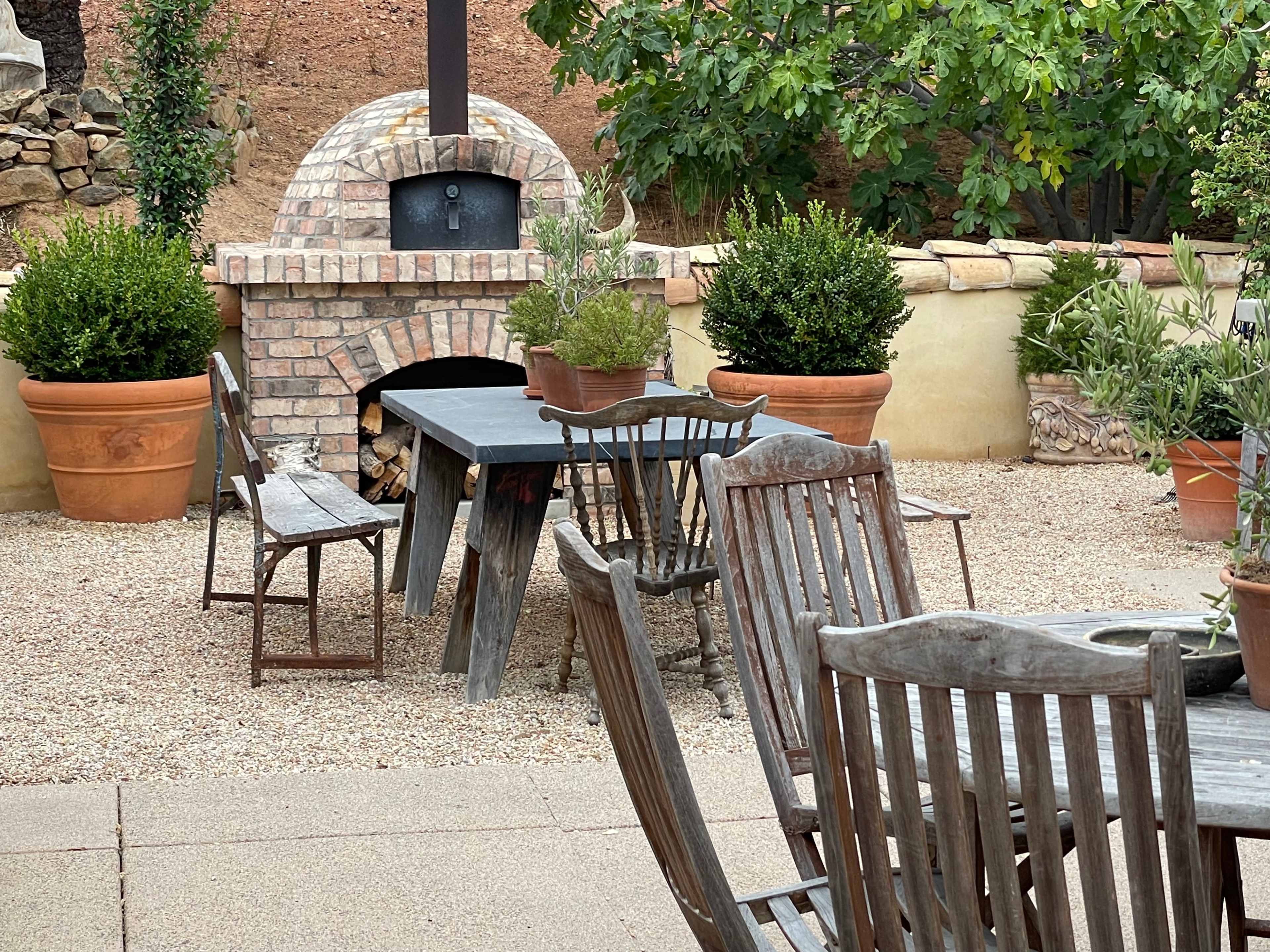 An outdoor patio area featuring a wooden table surrounded by chairs, with a brick oven and potted plants in the background.
