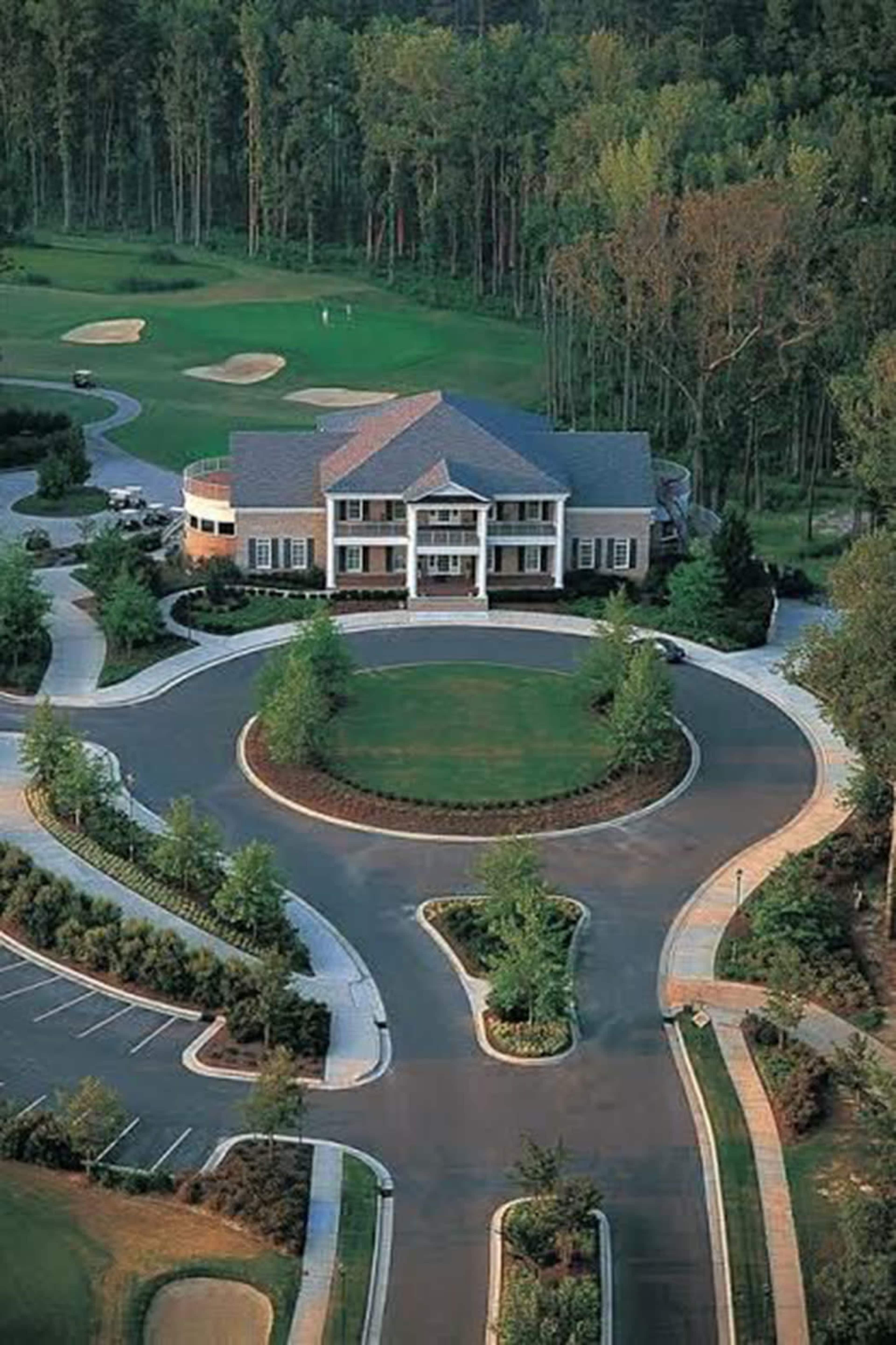 A large, elegant building with a circular driveway, surrounded by neatly landscaped greenery and a golf course in the background.