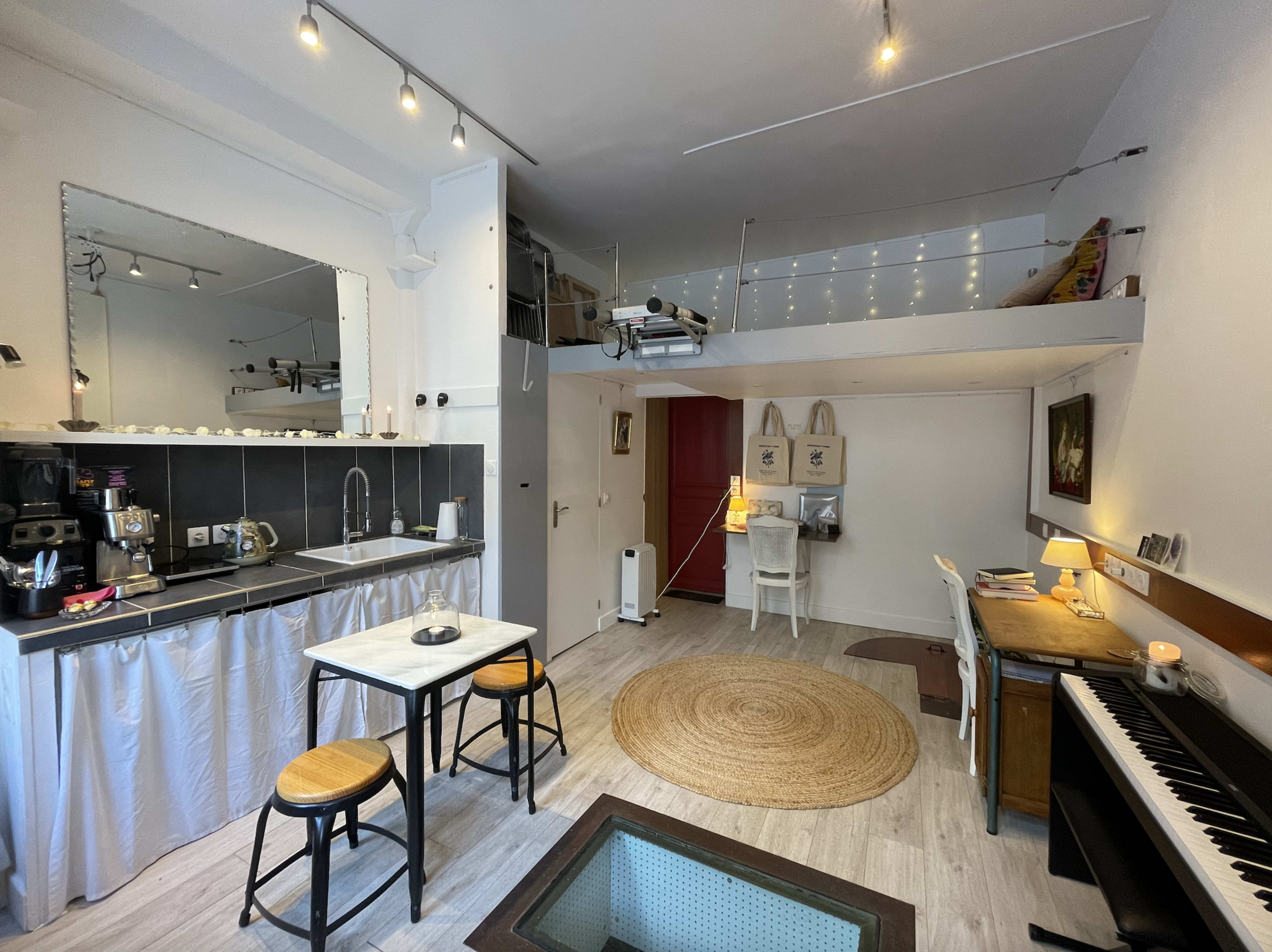 The image shows a small, modern studio apartment with a kitchen, dining area, and workspace, featuring a lofted sleeping area above.