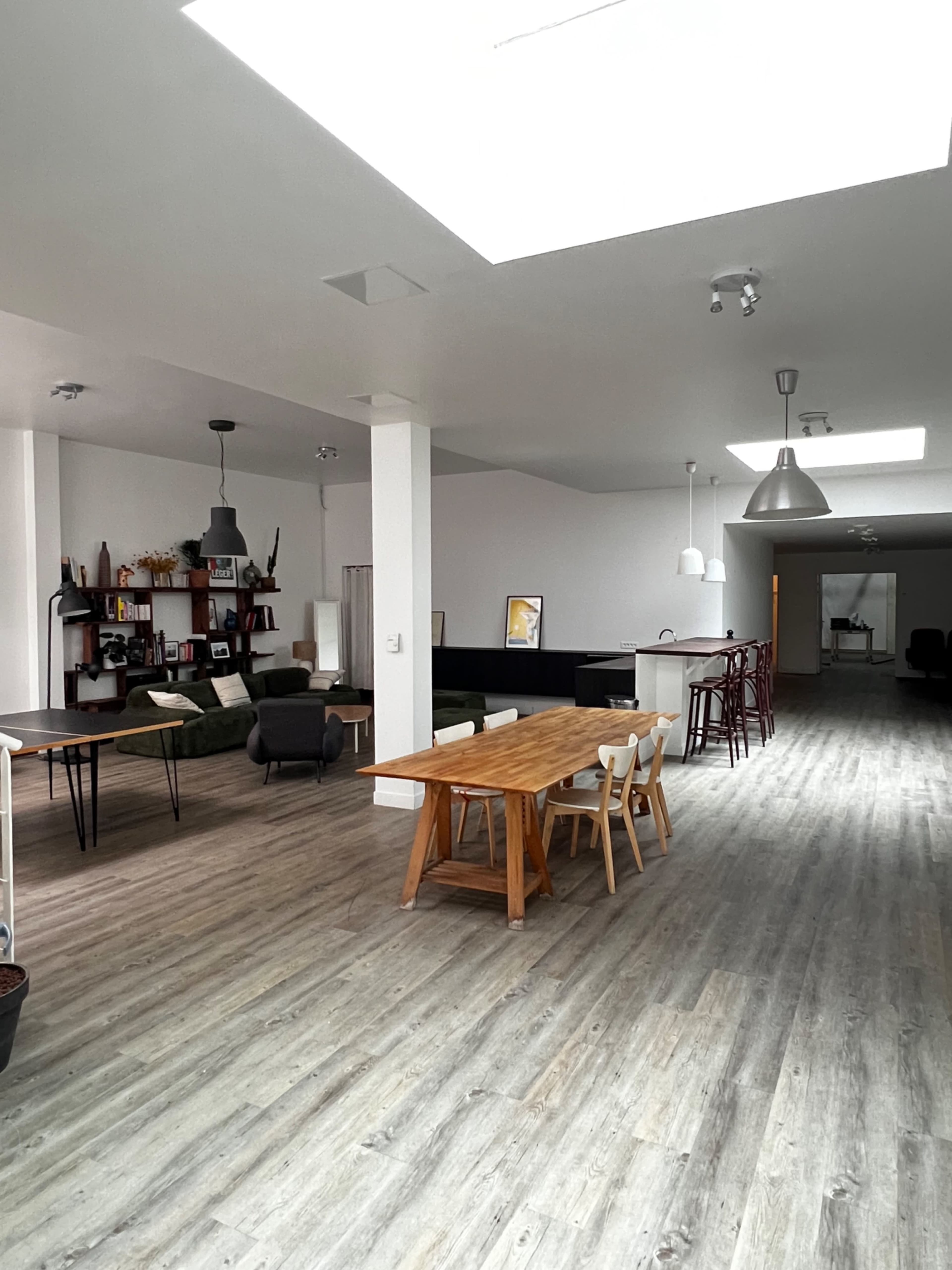 The image shows a spacious, modern office interior with large windows, a wooden dining table, various seating areas, and minimalist decor.