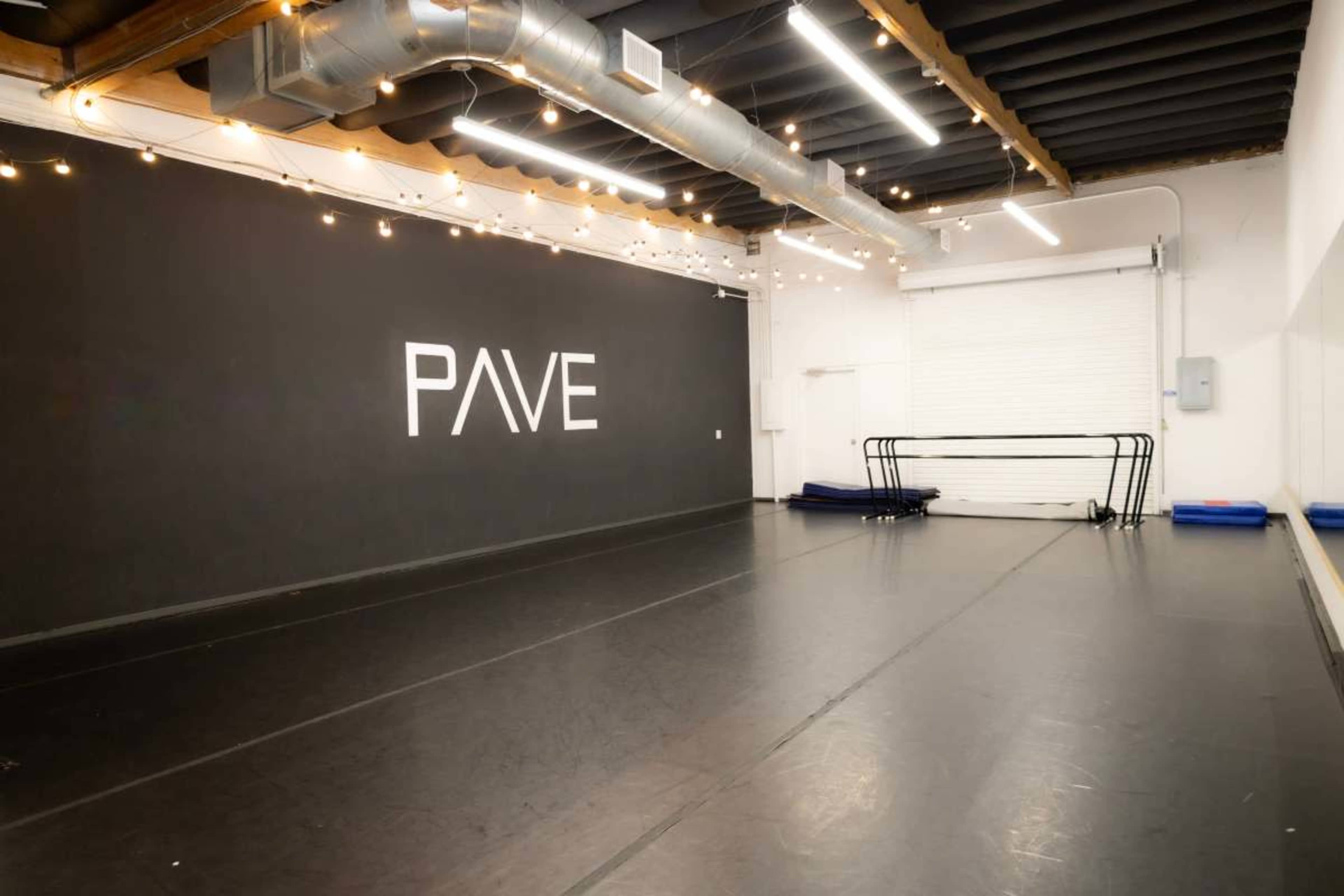The image shows a spacious dance studio featuring a black wall with the word "PAVE" illuminated in white, along with several exercise mats and a folding ballet barre.