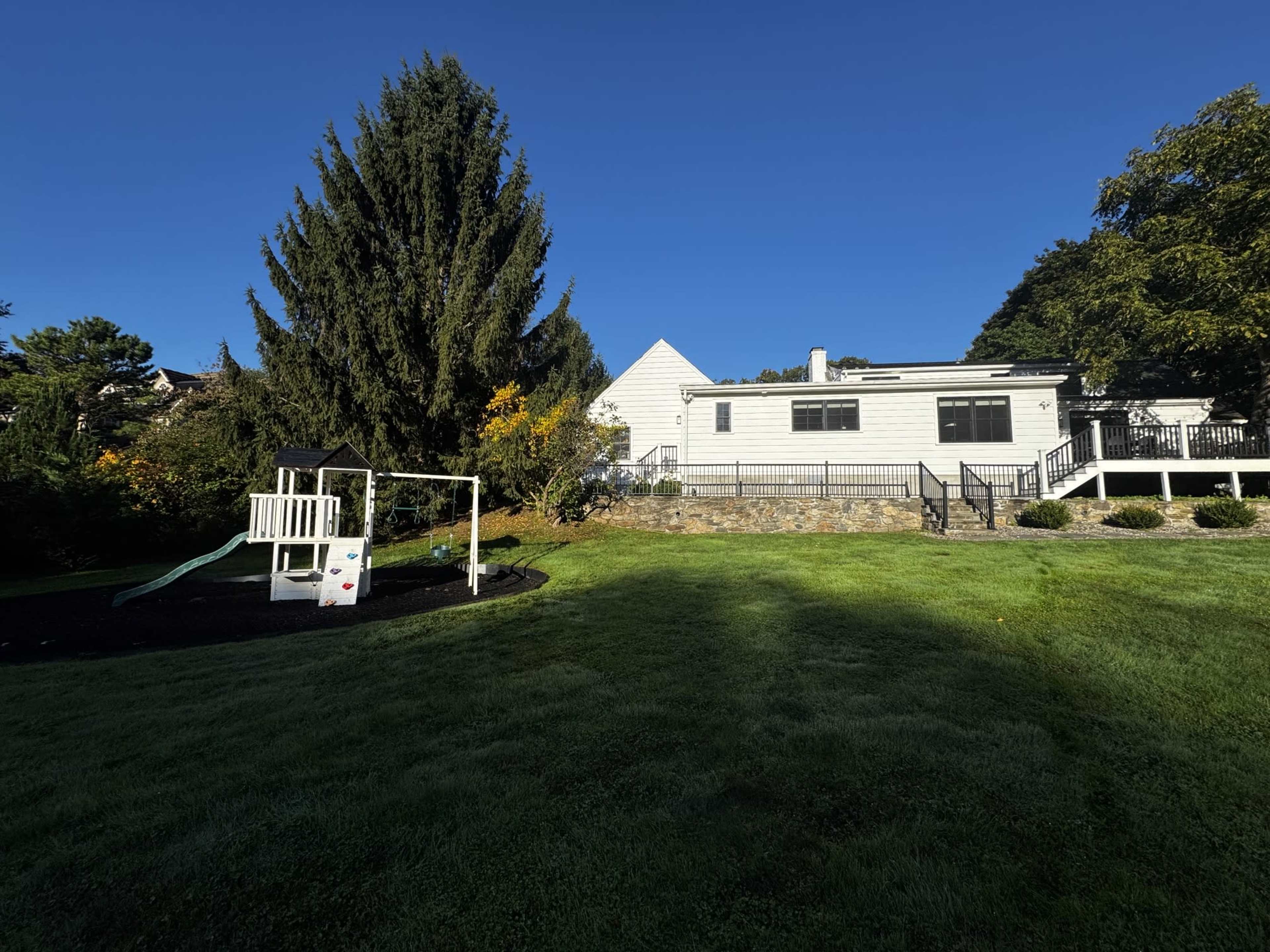 The image shows a backyard with a playground set on the left and a house featuring a large deck on the right, surrounded by trees and green grass.