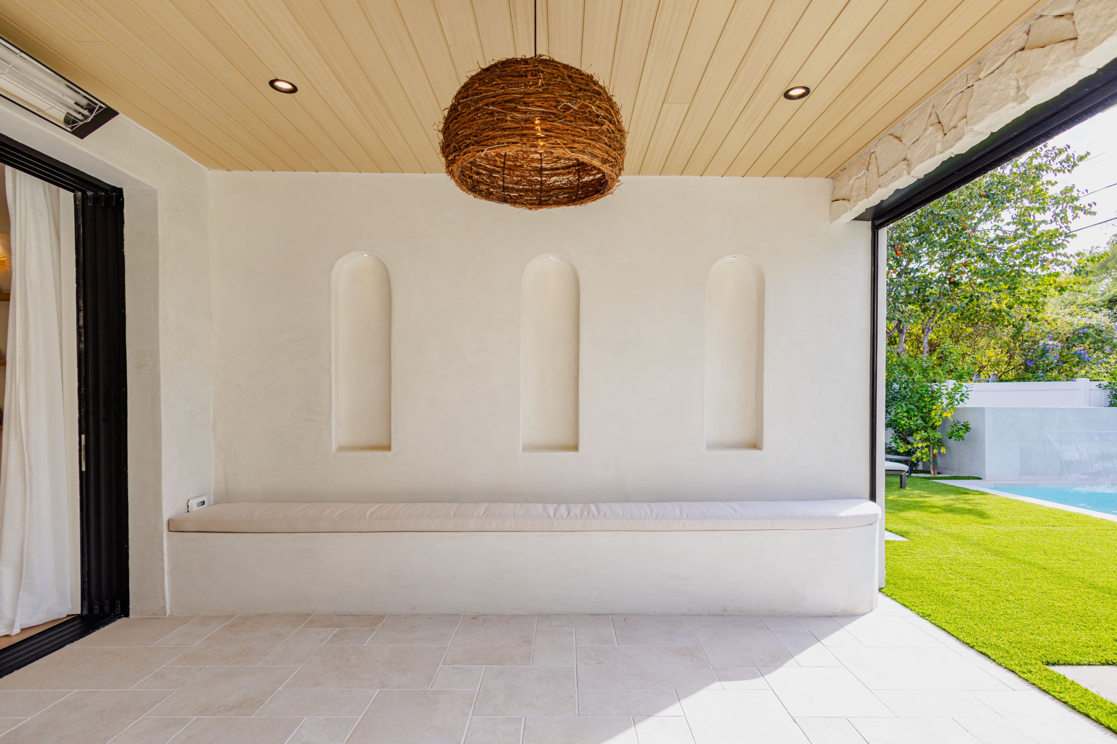 The image shows a bright indoor space featuring a smooth white wall with three recessed niches and a cushioned bench beneath a woven light fixture.