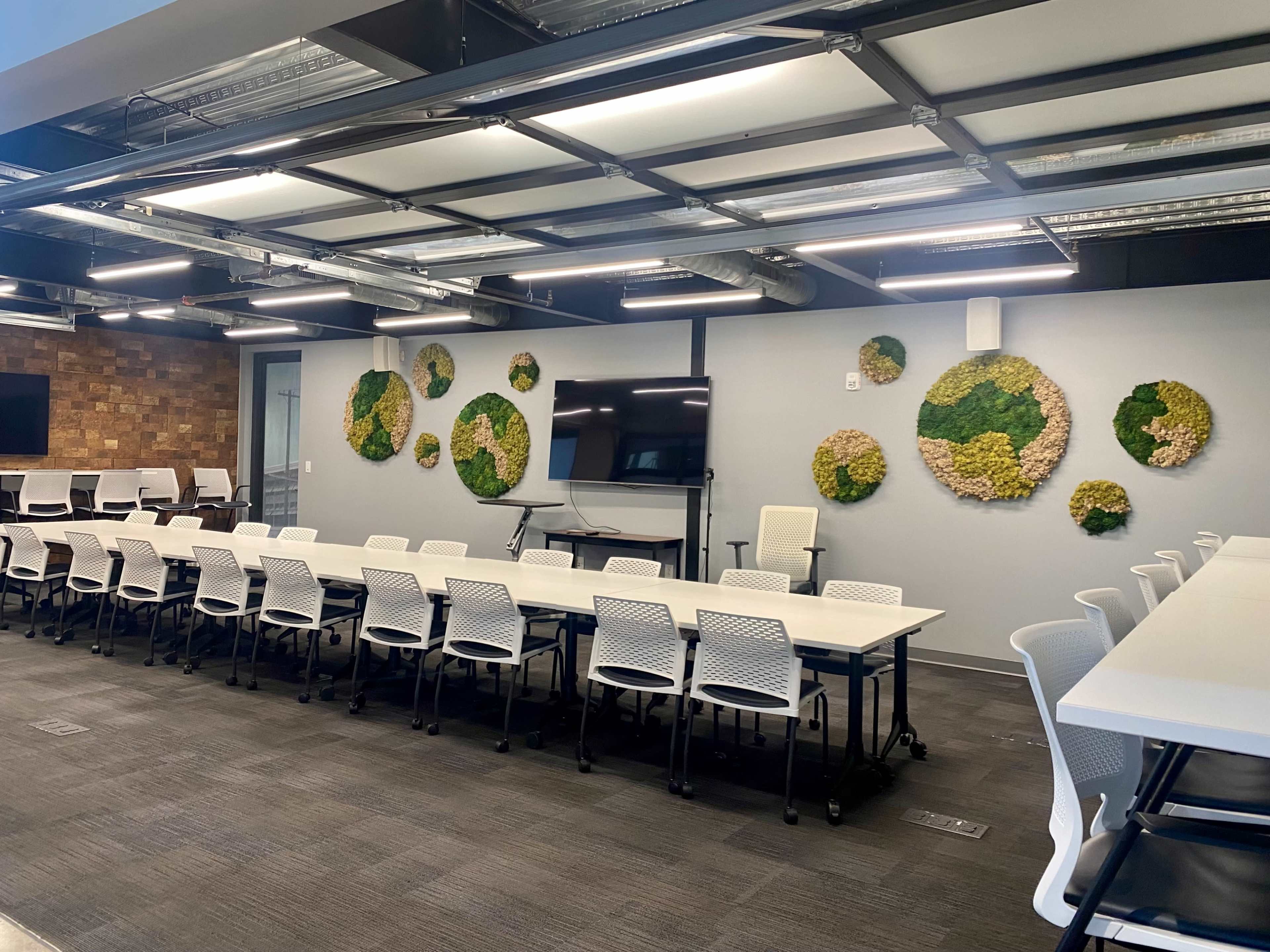 A modern conference room features long, white tables arranged in rows, with green moss wall art on the gray walls and a large screen at the front.