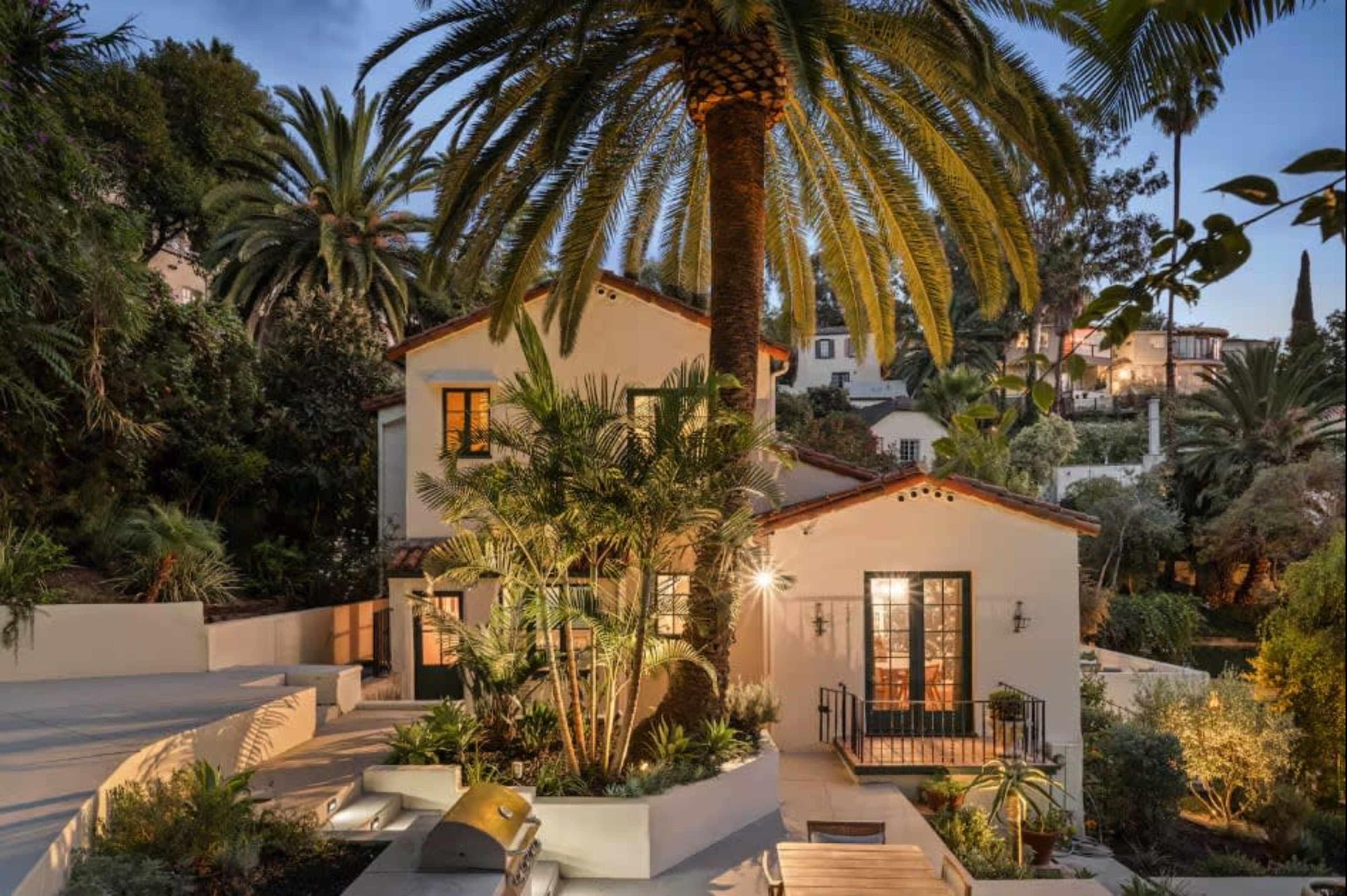 A two-story house with a white exterior and a lush garden is surrounded by palm trees and other greenery in a tranquil setting.