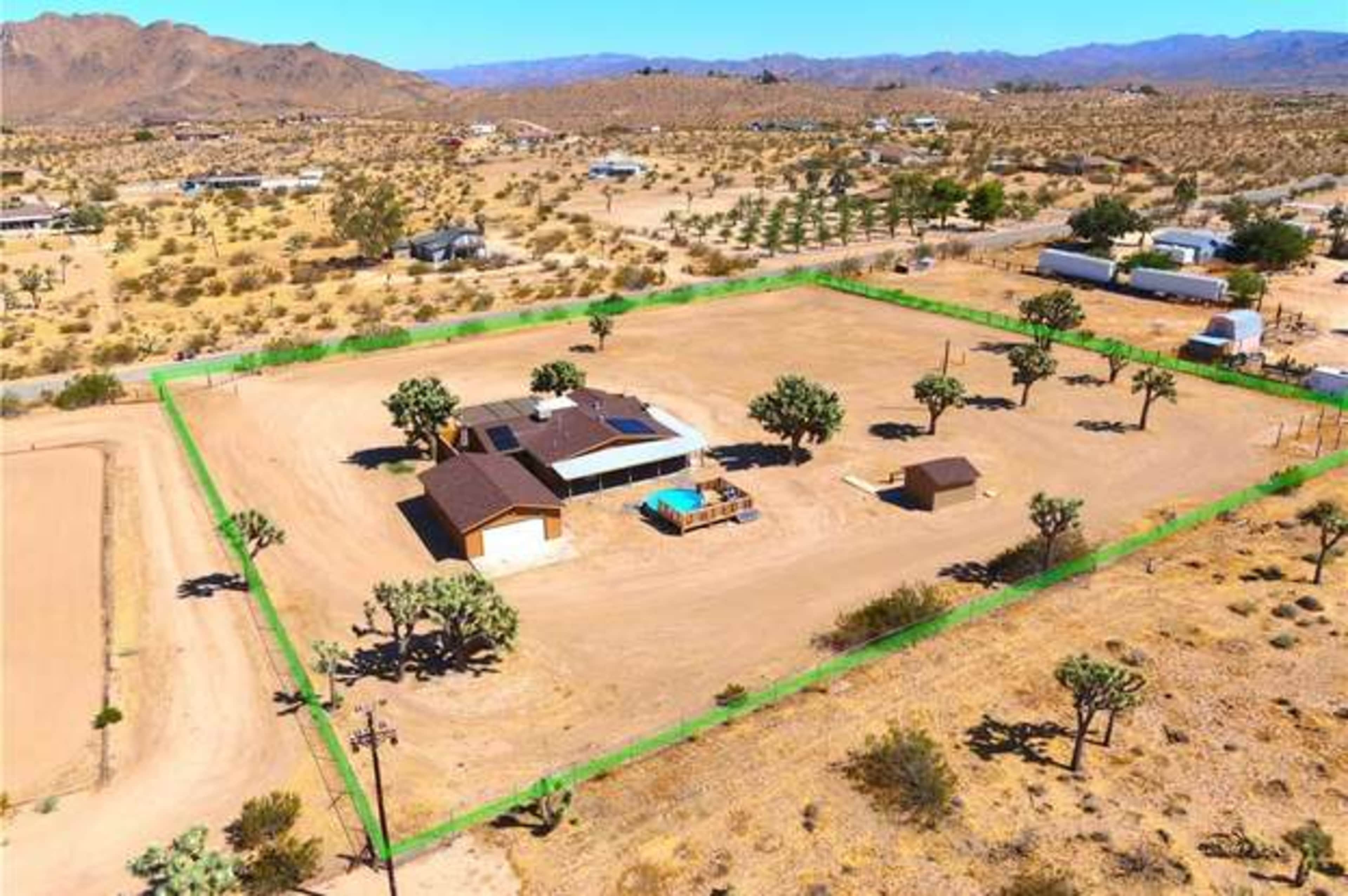 The image shows an aerial view of a single-story house surrounded by a large fenced area in a desert landscape, with sparse vegetation and distant mountains in the background.