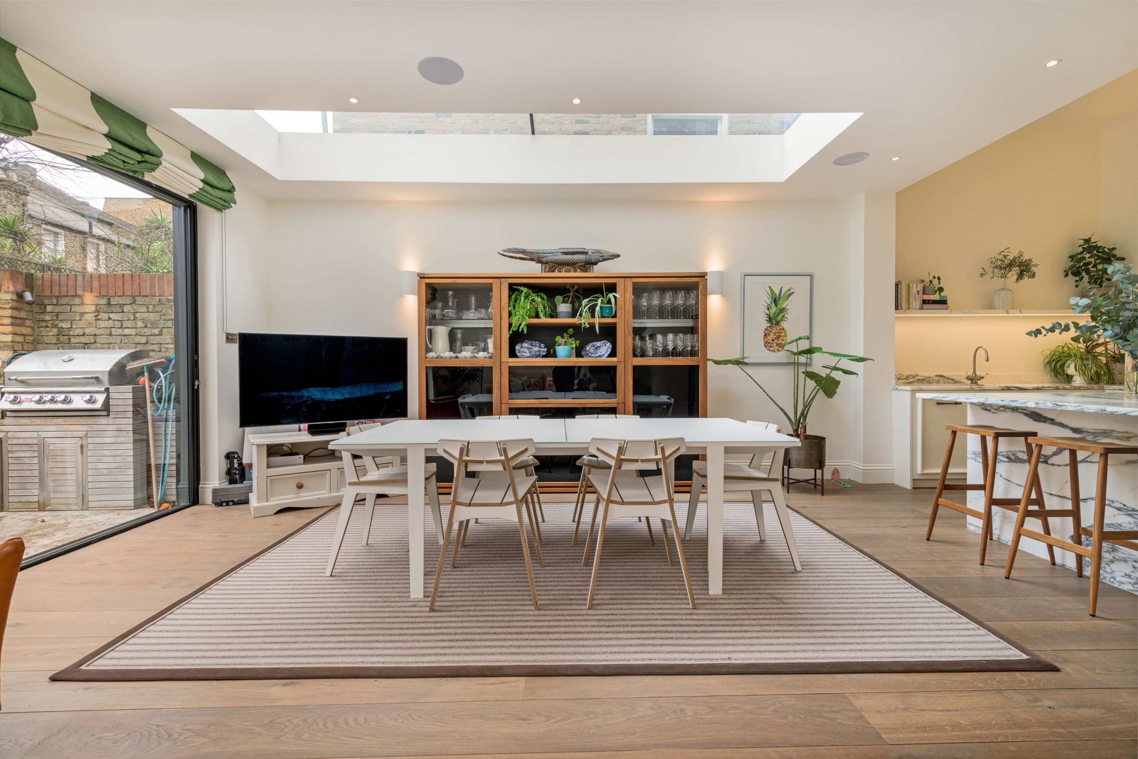 A modern dining area with a table and chairs, a television, a display cabinet, and a kitchenette illuminated by natural light from a skylight.