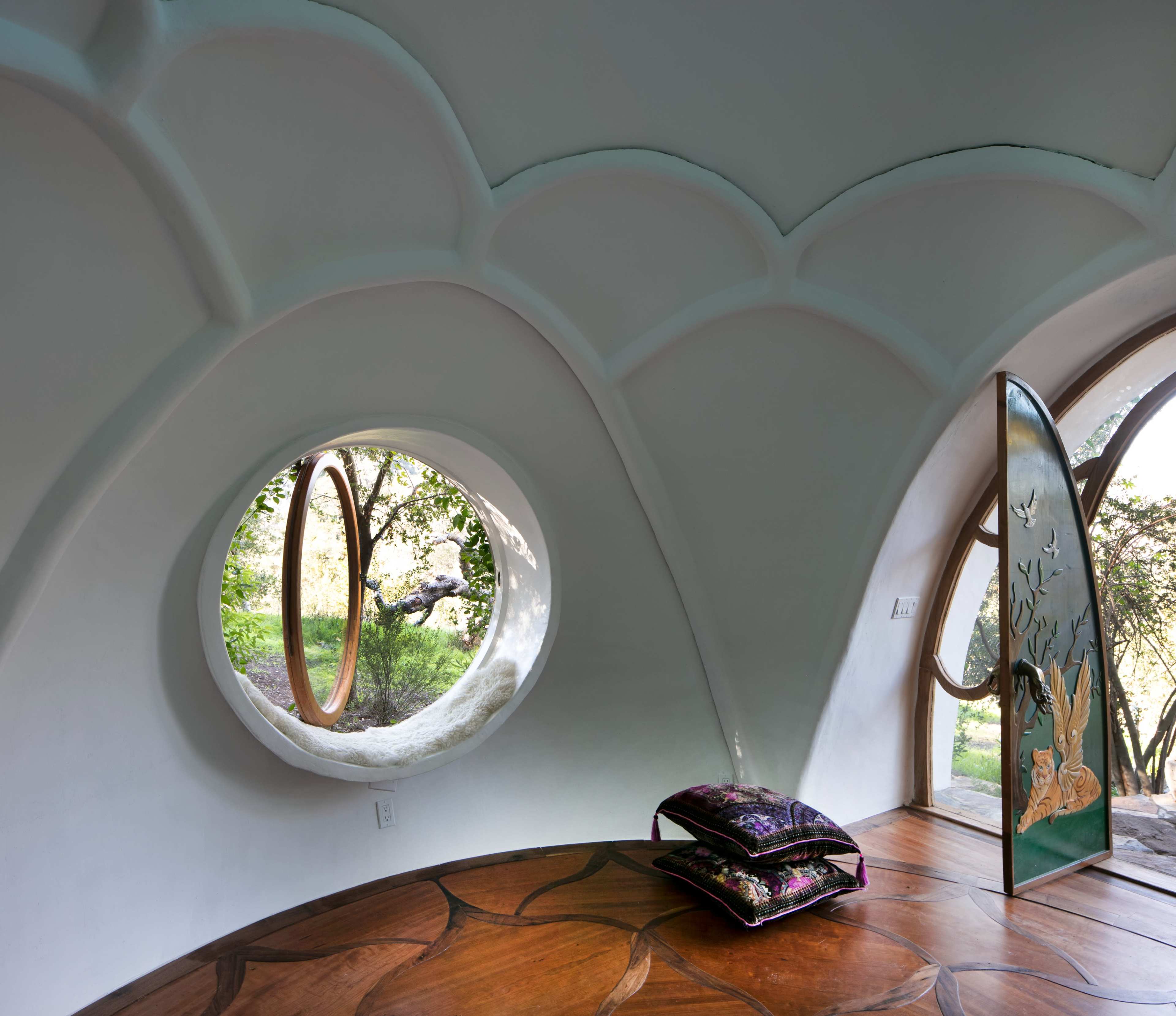 The interior of a round room features a circular window, a wooden floor with a unique pattern, and a decorative door that opens to a green outdoor view.