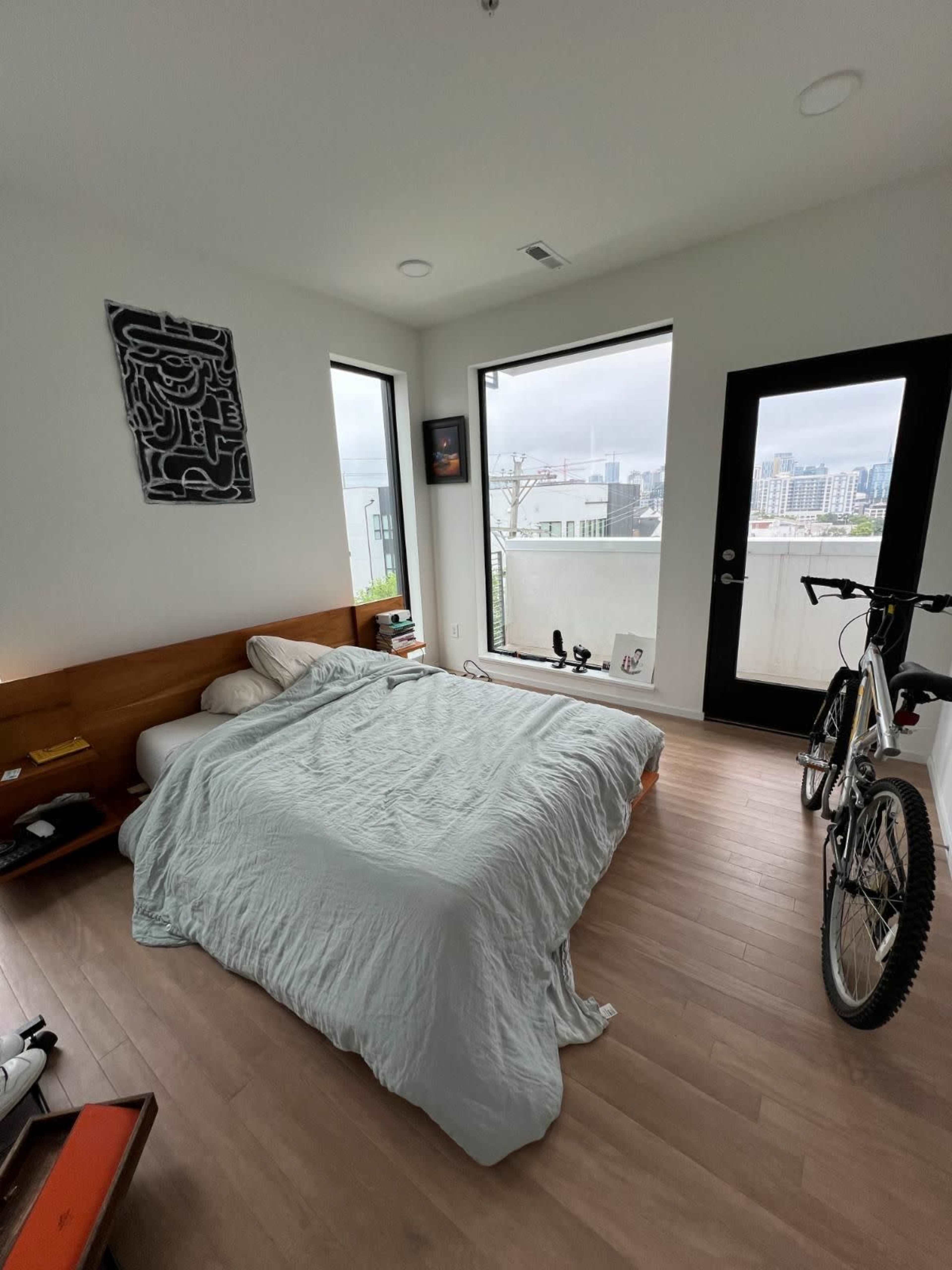 The bedroom features a bed with a light-colored quilt, large windows overlooking a cityscape, and a bicycle parked next to the door.
