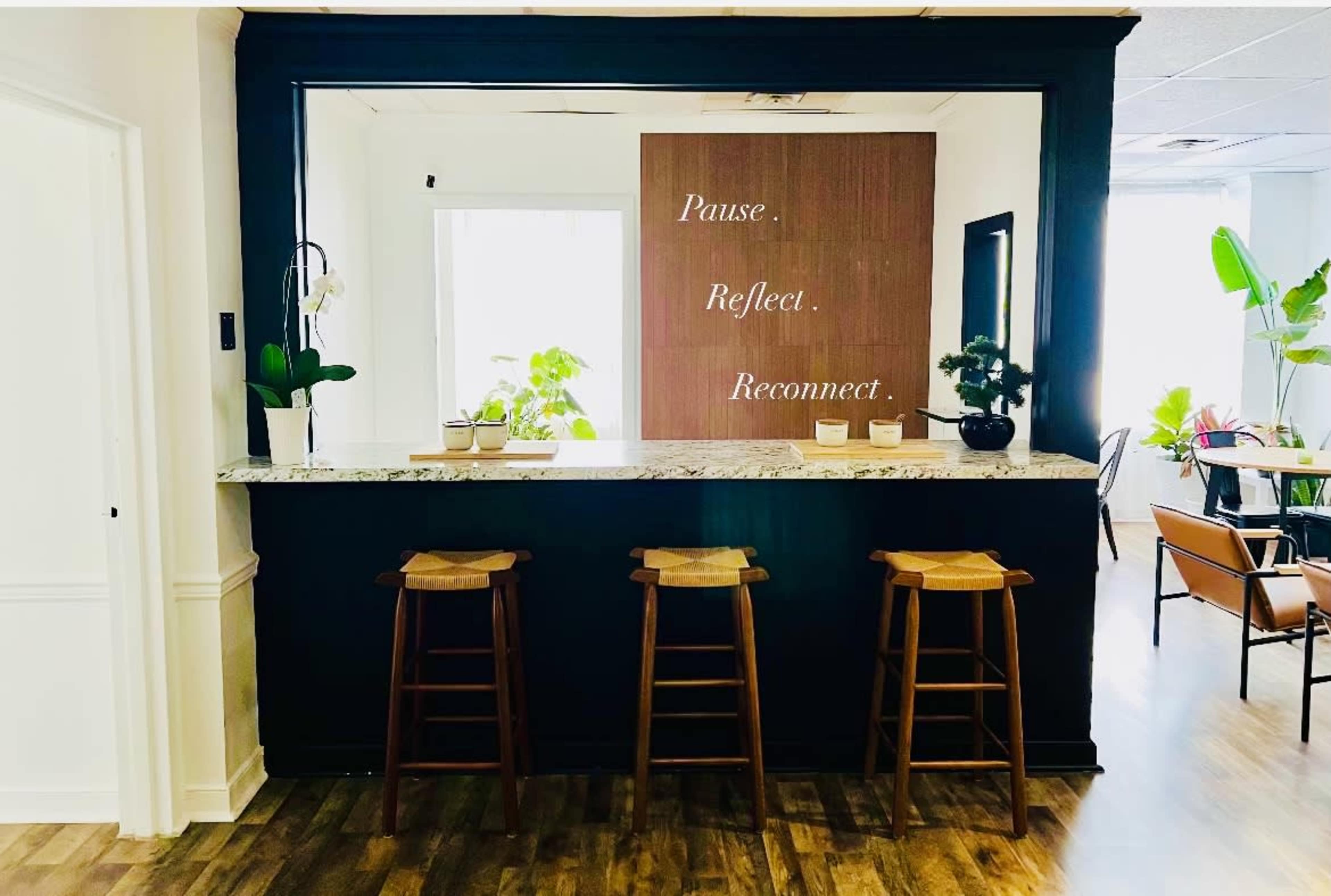 A minimalist reception area features a granite counter with three wooden stools and a wall displaying the words "Pause. Reflect. Reconnect."