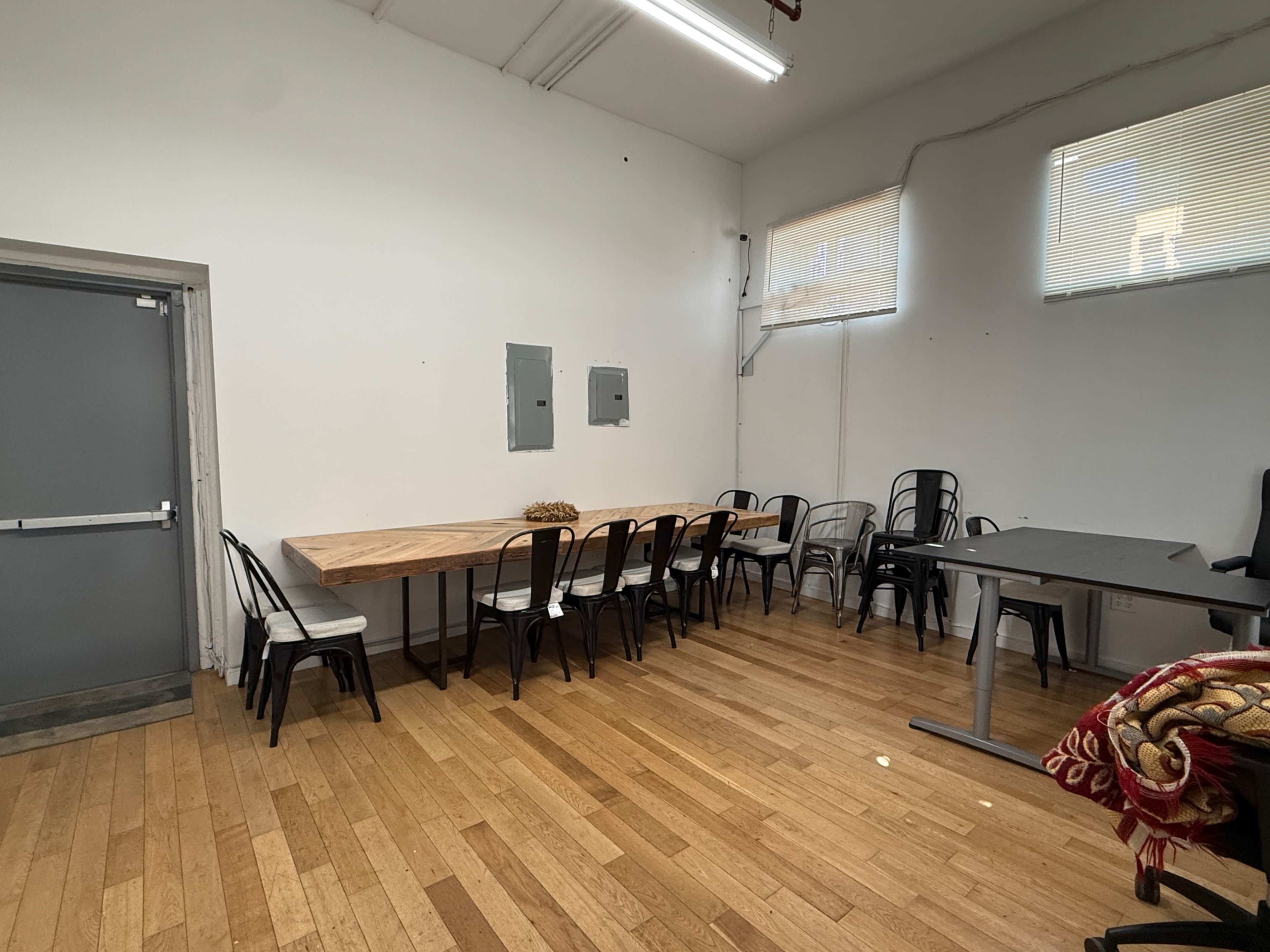 The image shows a large room with a wooden table surrounded by metal and plastic chairs, along with an office desk in the corner.