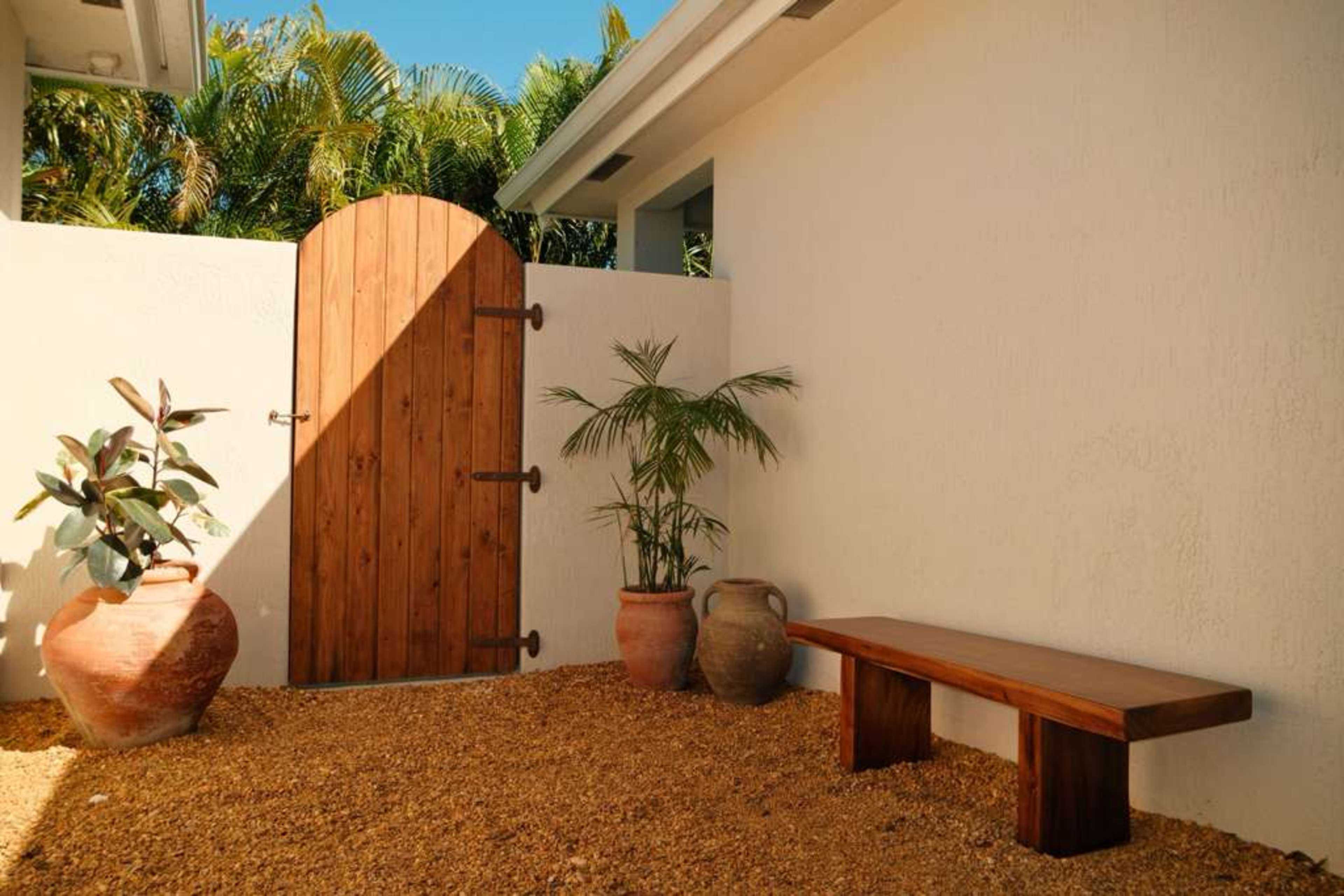 The image shows a small, enclosed outdoor space featuring a wooden bench, a few clay pots, and a wooden door against a white wall, surrounded by gravel and green plants.