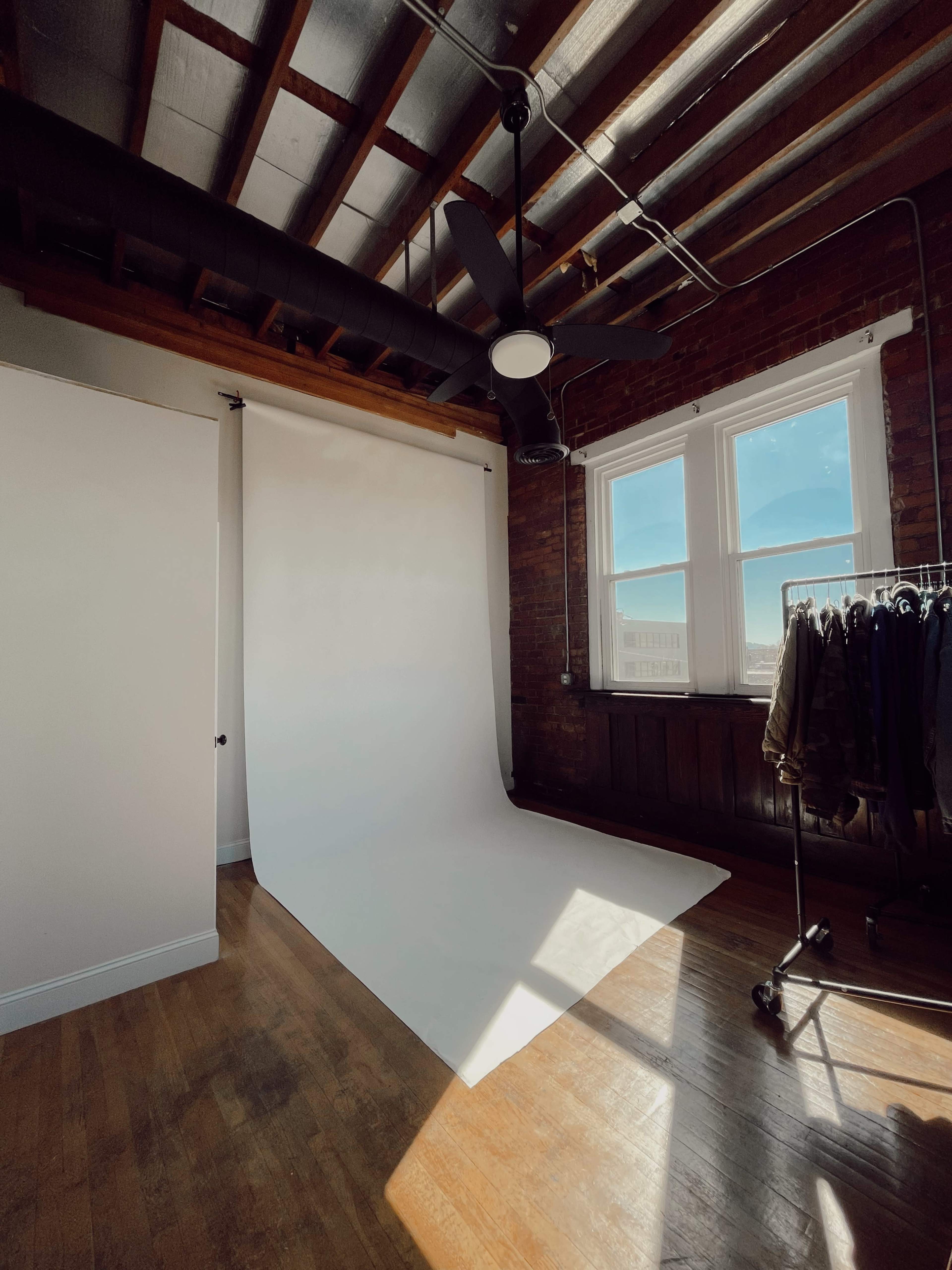 A spacious, well-lit room with a white backdrop, large windows, and a clothing rack against a brick wall.