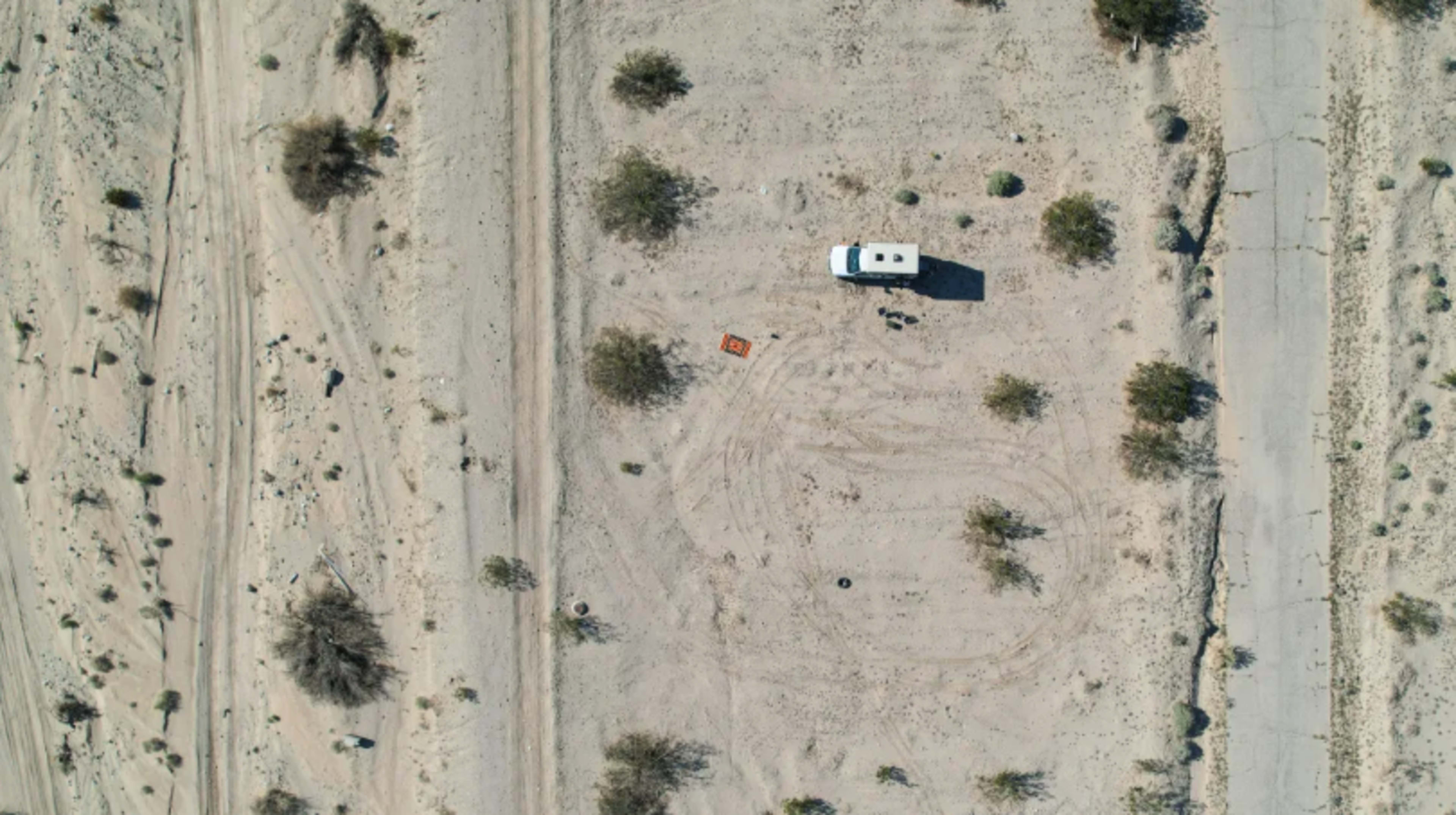 An aerial view shows a white RV parked on a barren, sandy landscape with sparse vegetation.