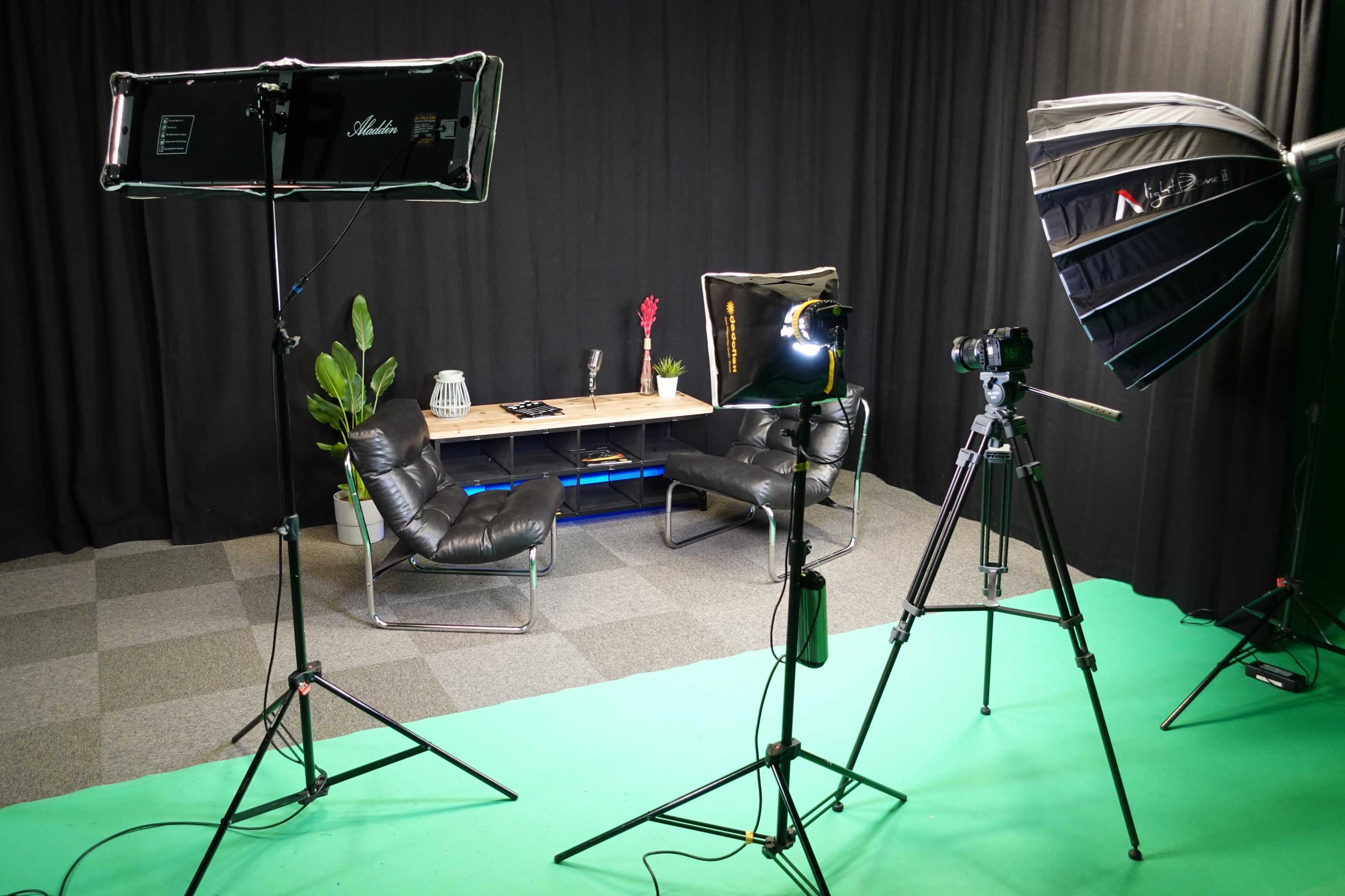 A film studio setup with lighting equipment, a camera on a tripod, and a seating area, all arranged on a green screen floor.