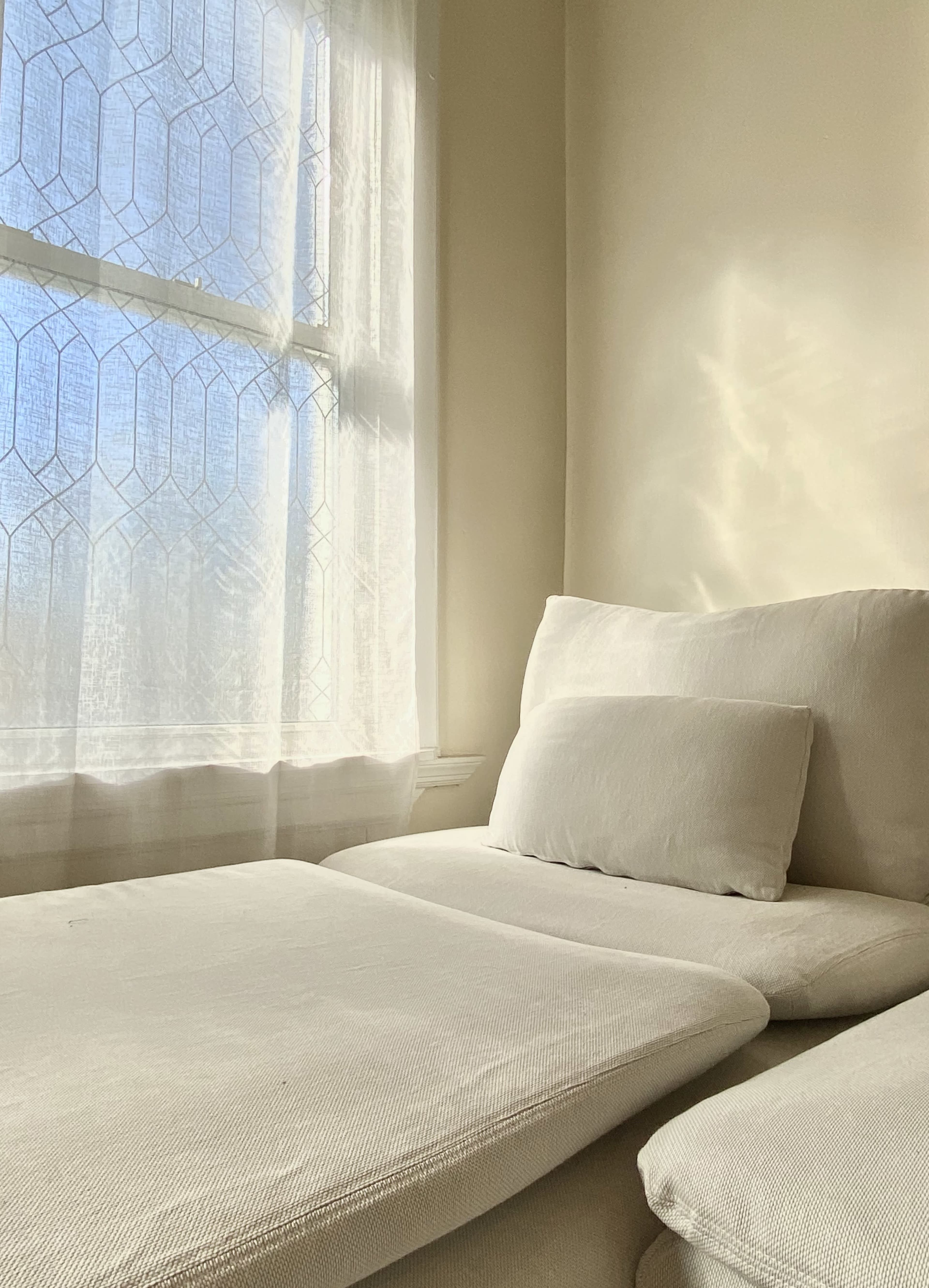 A light-colored upholstered chaise lounge sits by a window adorned with sheer curtains, allowing sunlight to filter in.