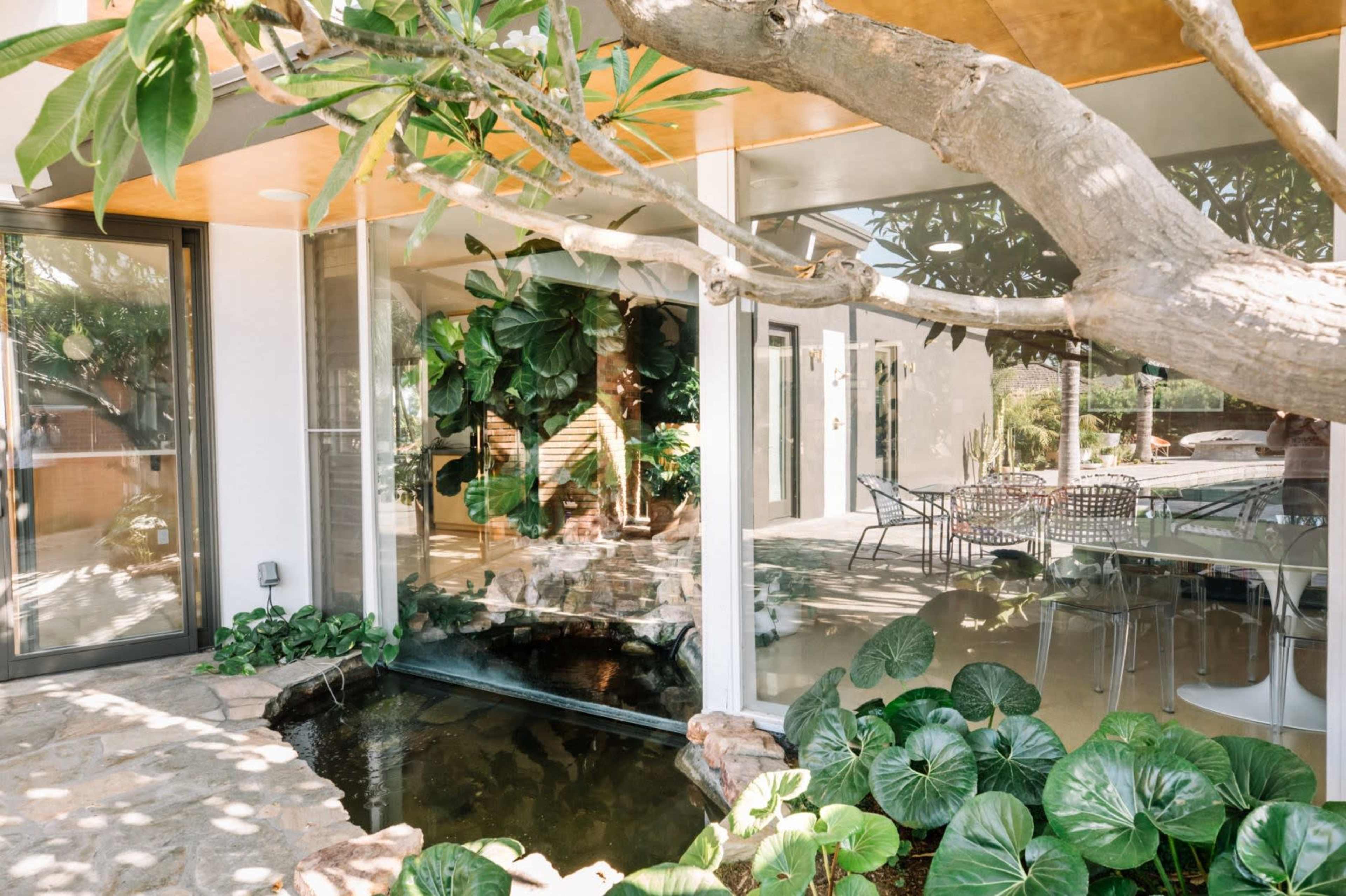 The image features a contemporary house with large glass walls surrounded by lush greenery and a small pond.