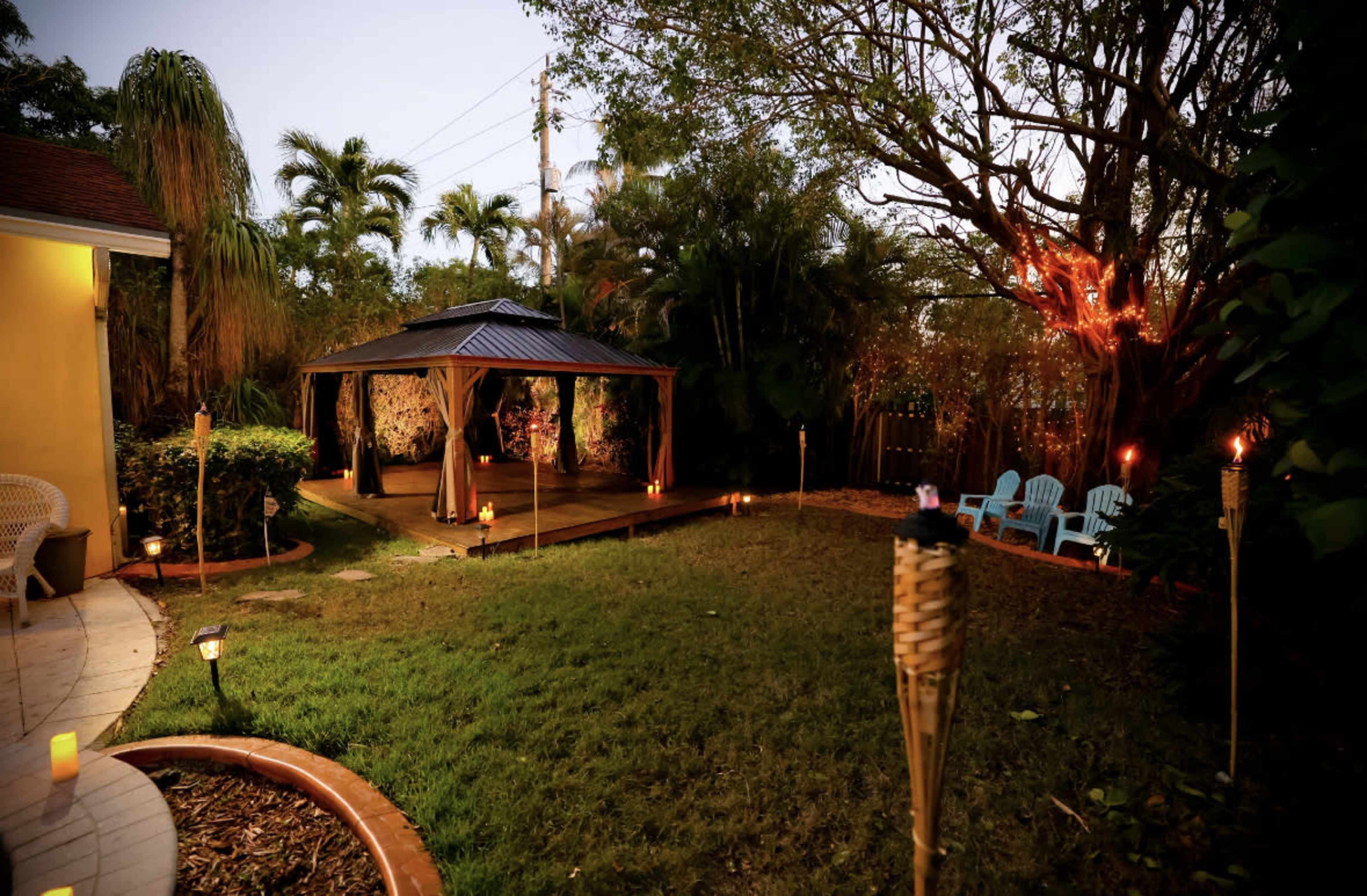 A landscaped backyard features a gazebo surrounded by tiki torches and chairs, with palm trees in the background.