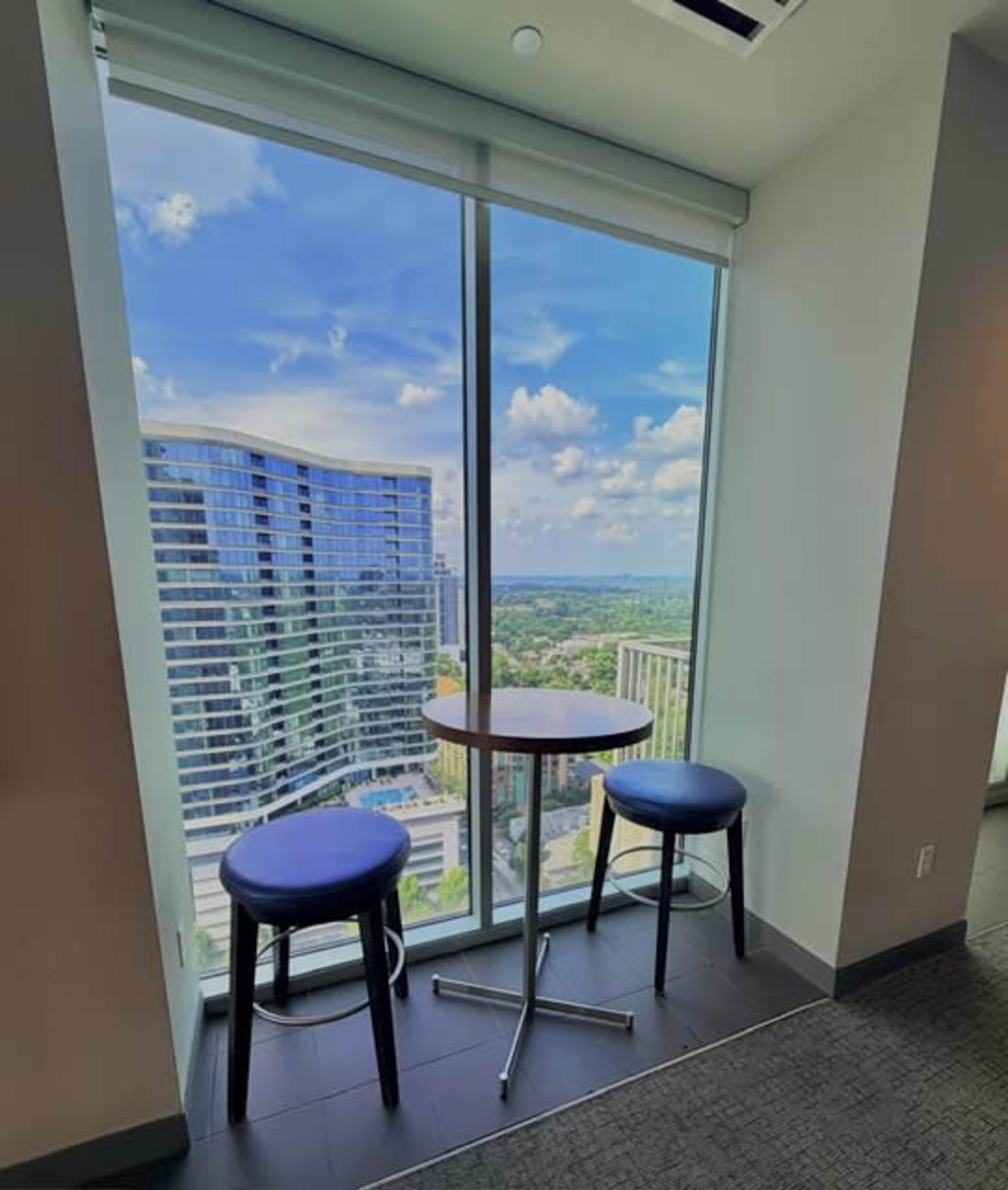 A small round table with two chairs sits by a large window, offering a view of nearby high-rise buildings and a clear blue sky.