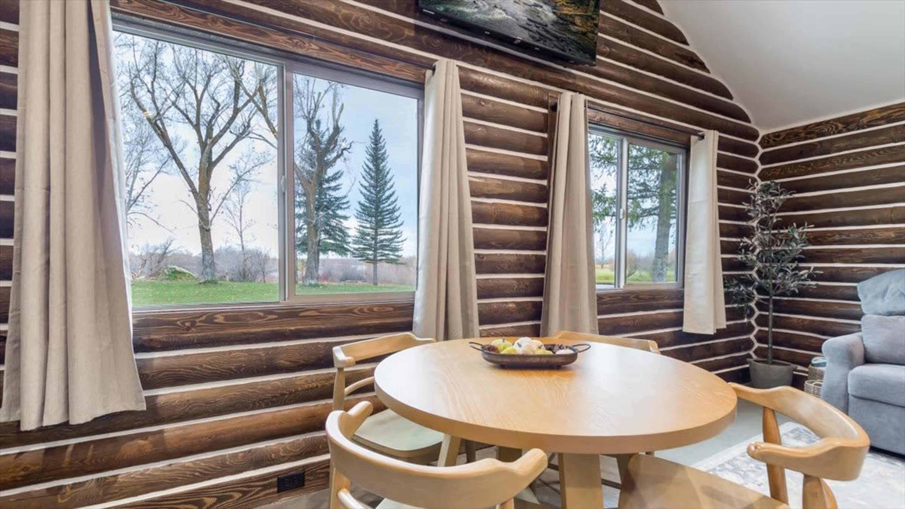 The image shows a cozy log cabin interior with a round wooden table, two chairs, and large windows revealing trees outside.