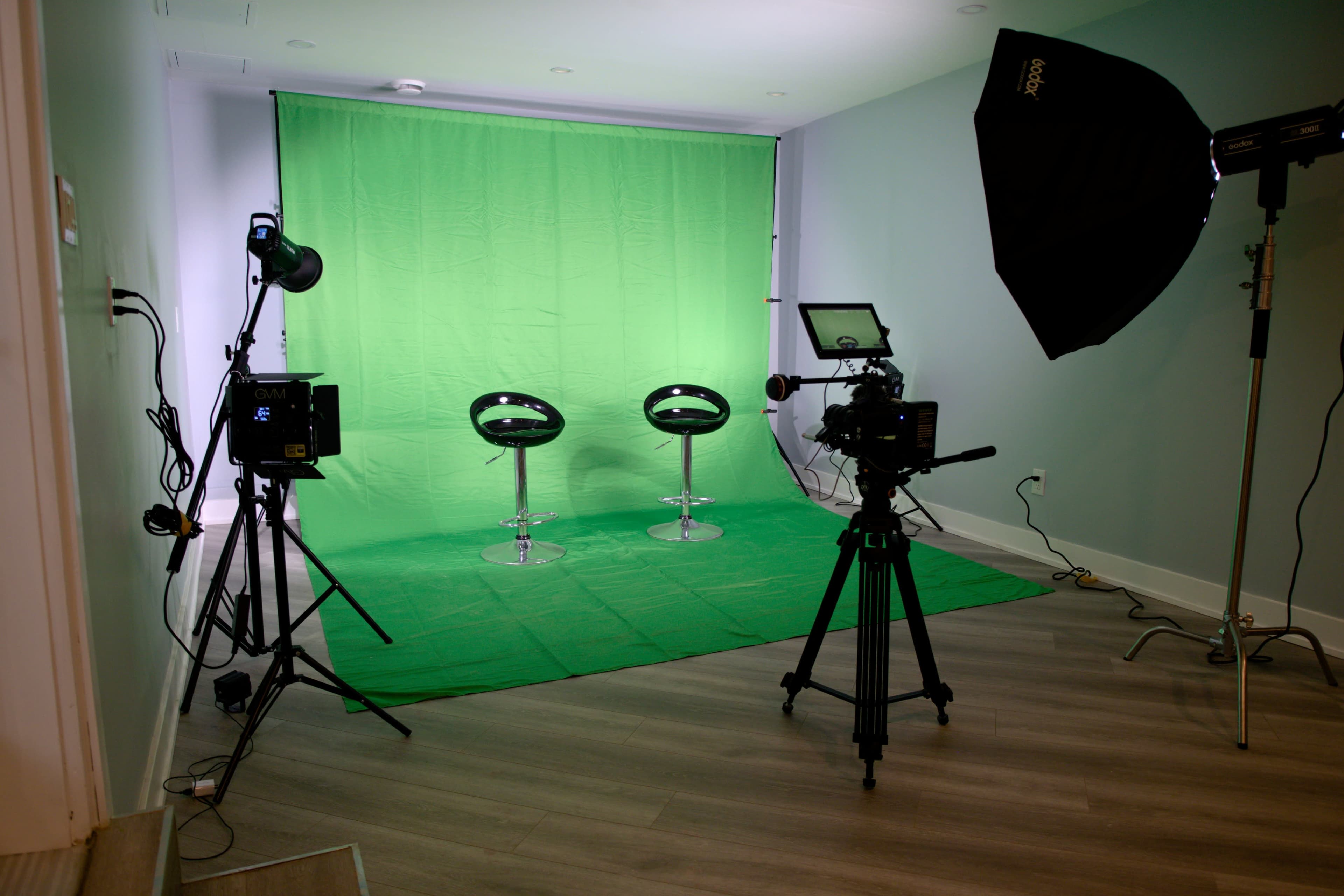 A photography studio with two black chairs on a green backdrop, illuminated by various lights and equipped with a camera on a tripod.