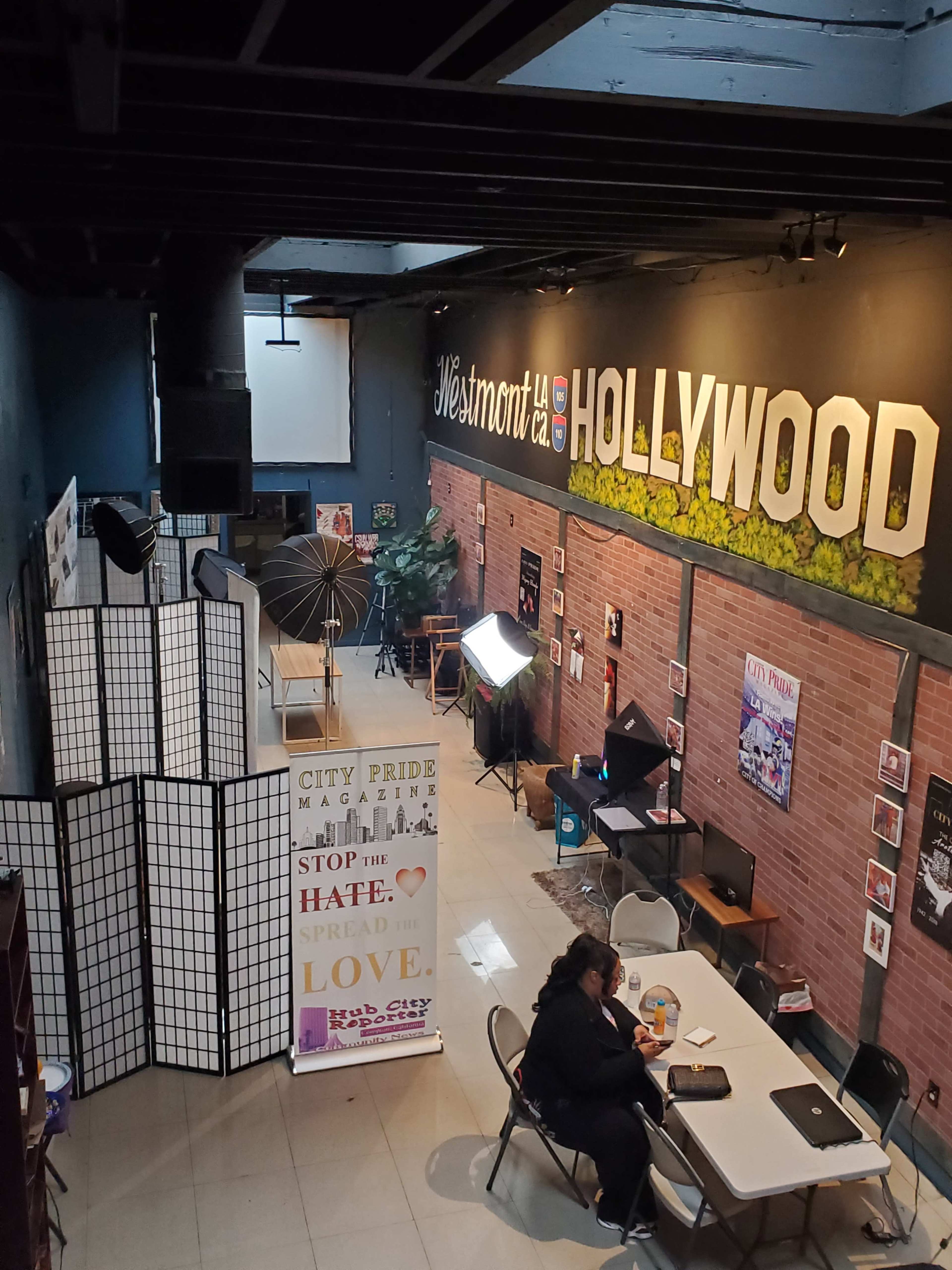 The image shows the interior of a community space in Westmont, West Hollywood, featuring decorative walls, tables, and promotional displays for "City Pride Magazine."