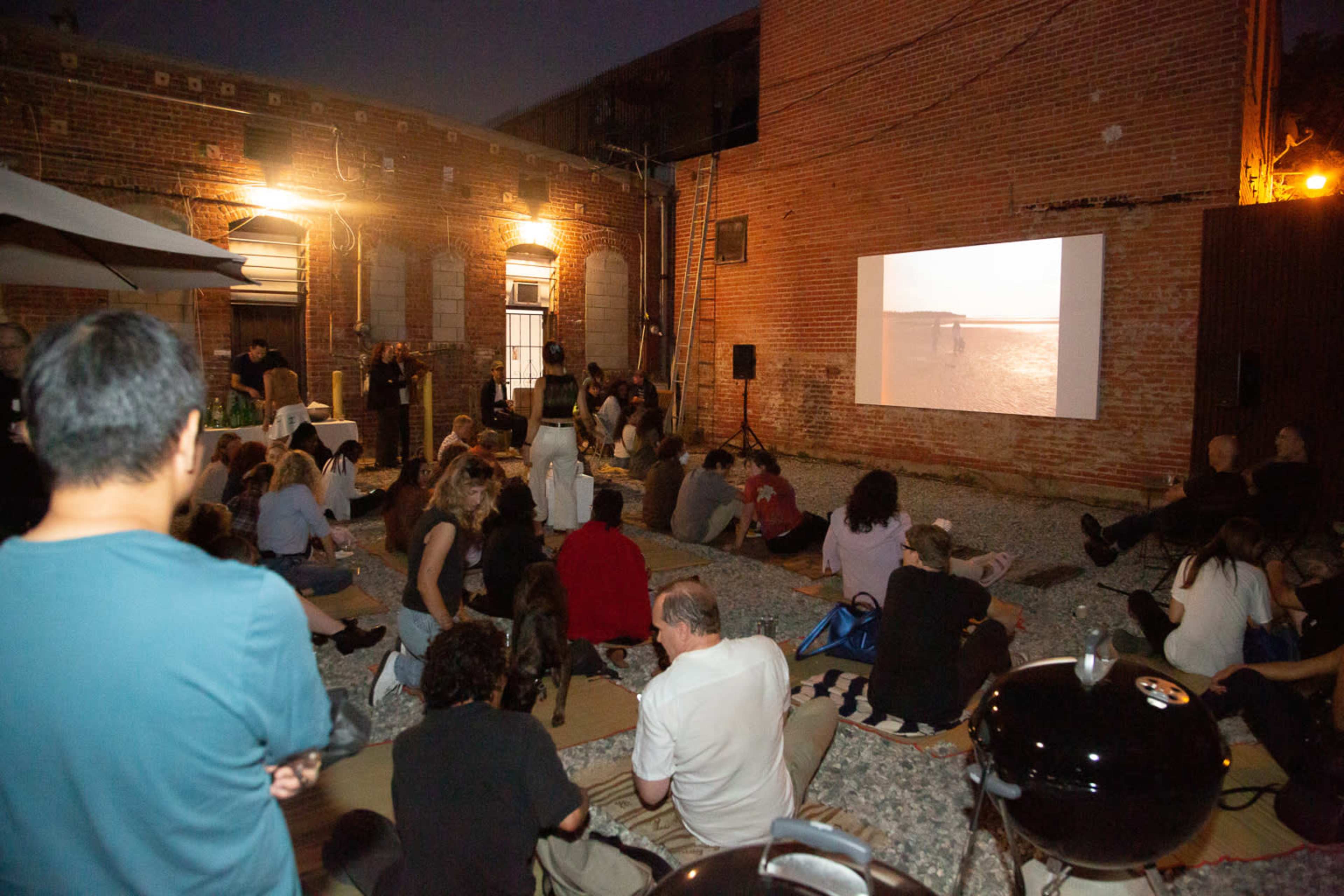 A large group of people sits on the ground in an outdoor space, watching a film projected on a wall.
