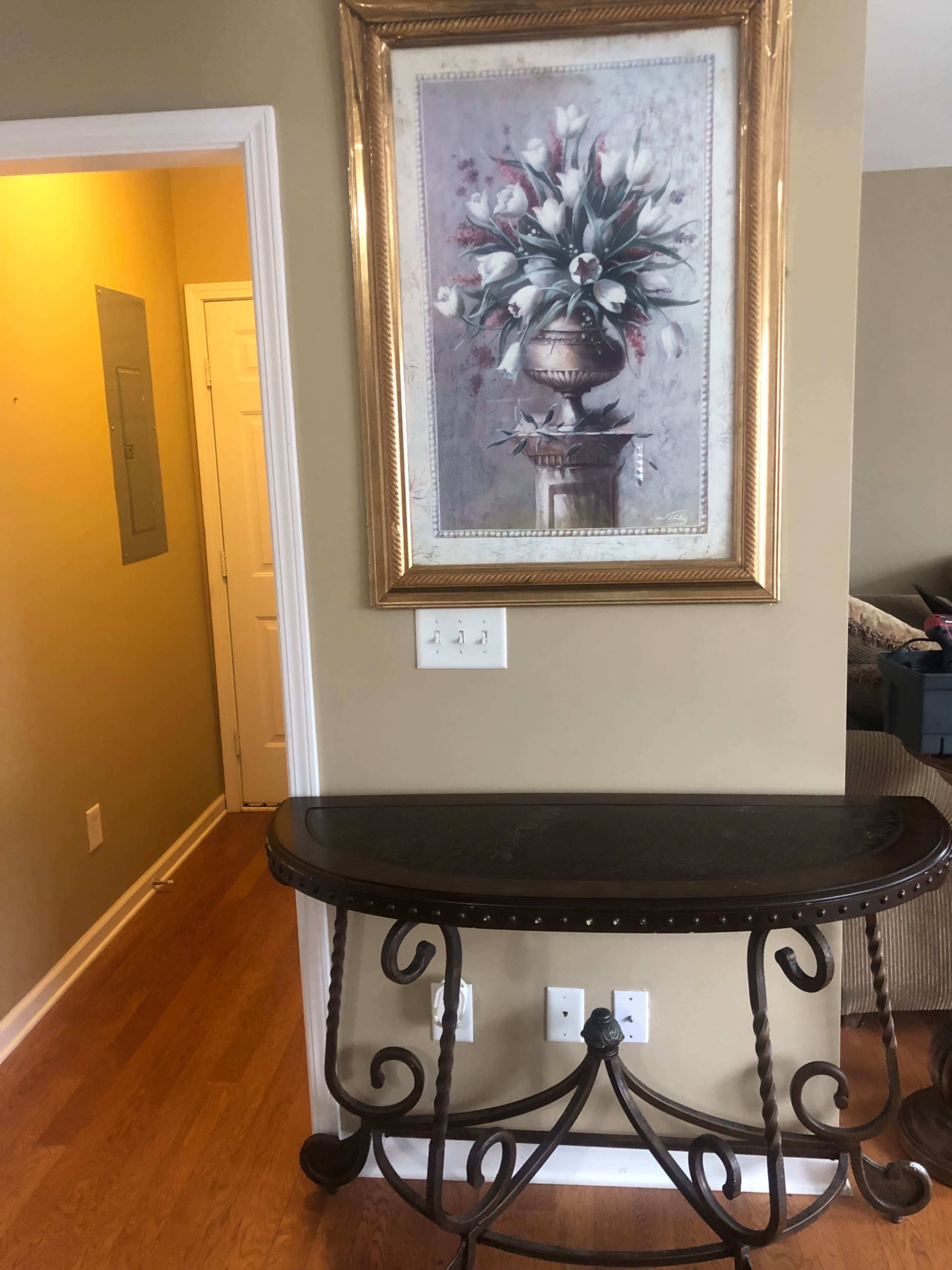 A console table against a wall with a framed floral painting above it, leading to a hallway on the left.