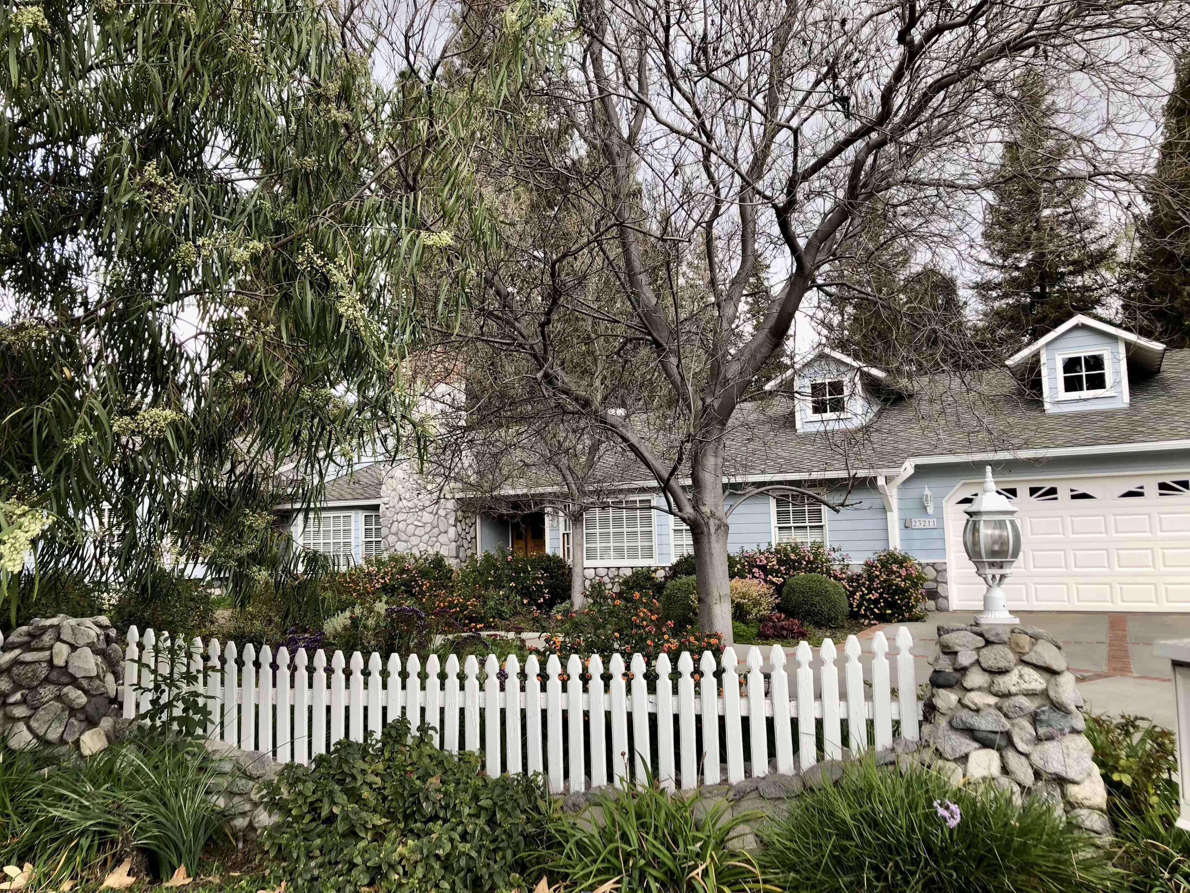 A house with a light blue exterior, surrounded by a white picket fence and various shrubs and trees, is visible in the scene.