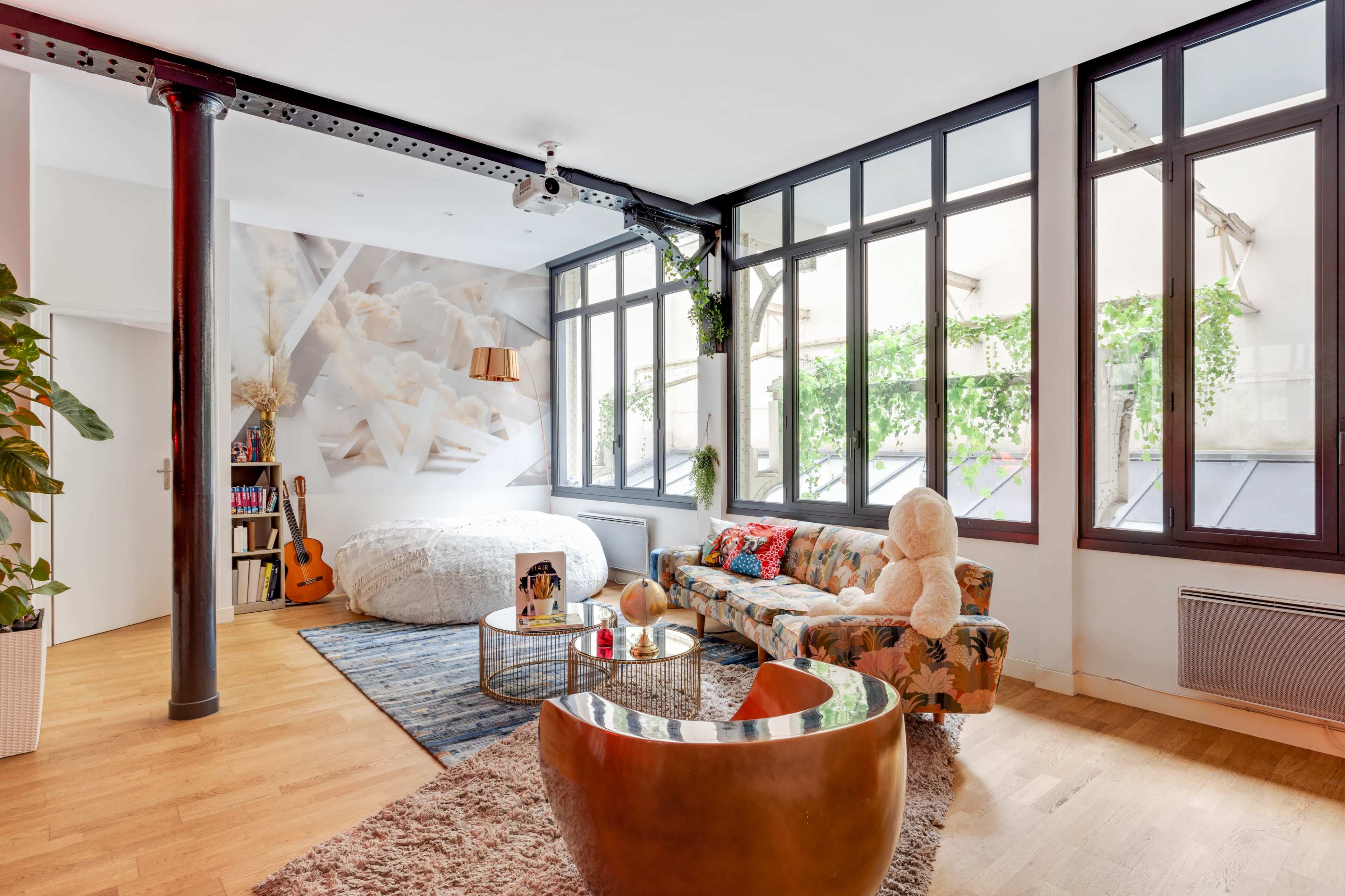 The image depicts a modern living space with large windows, a round bed, a floral sofa, and a wooden coffee table, complemented by decorative plants.