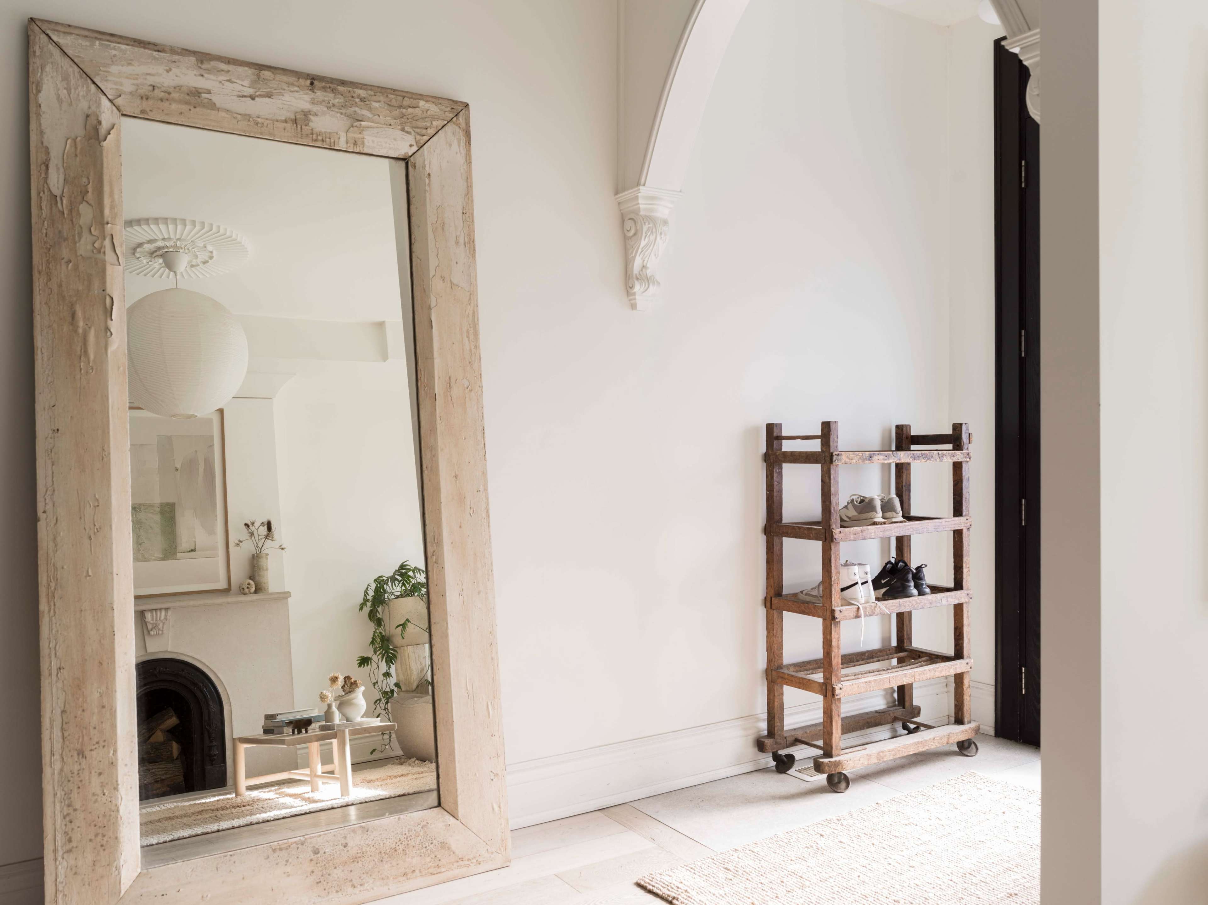 A large, weathered mirror leans against a white wall beside a wooden shoe rack with several pairs of shoes.