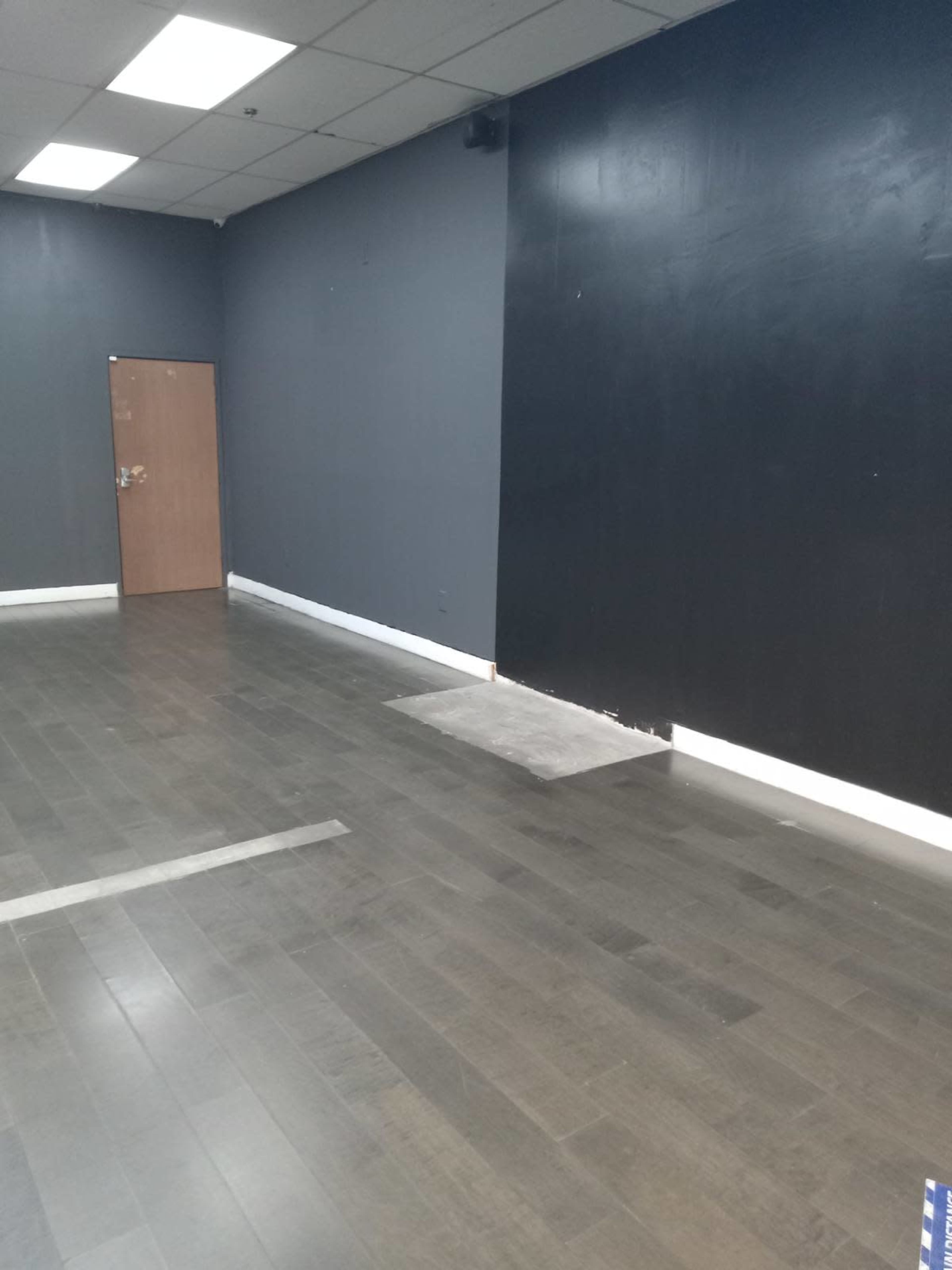 The image depicts an empty room with gray walls, a wooden floor, and a closed wooden door on one side.