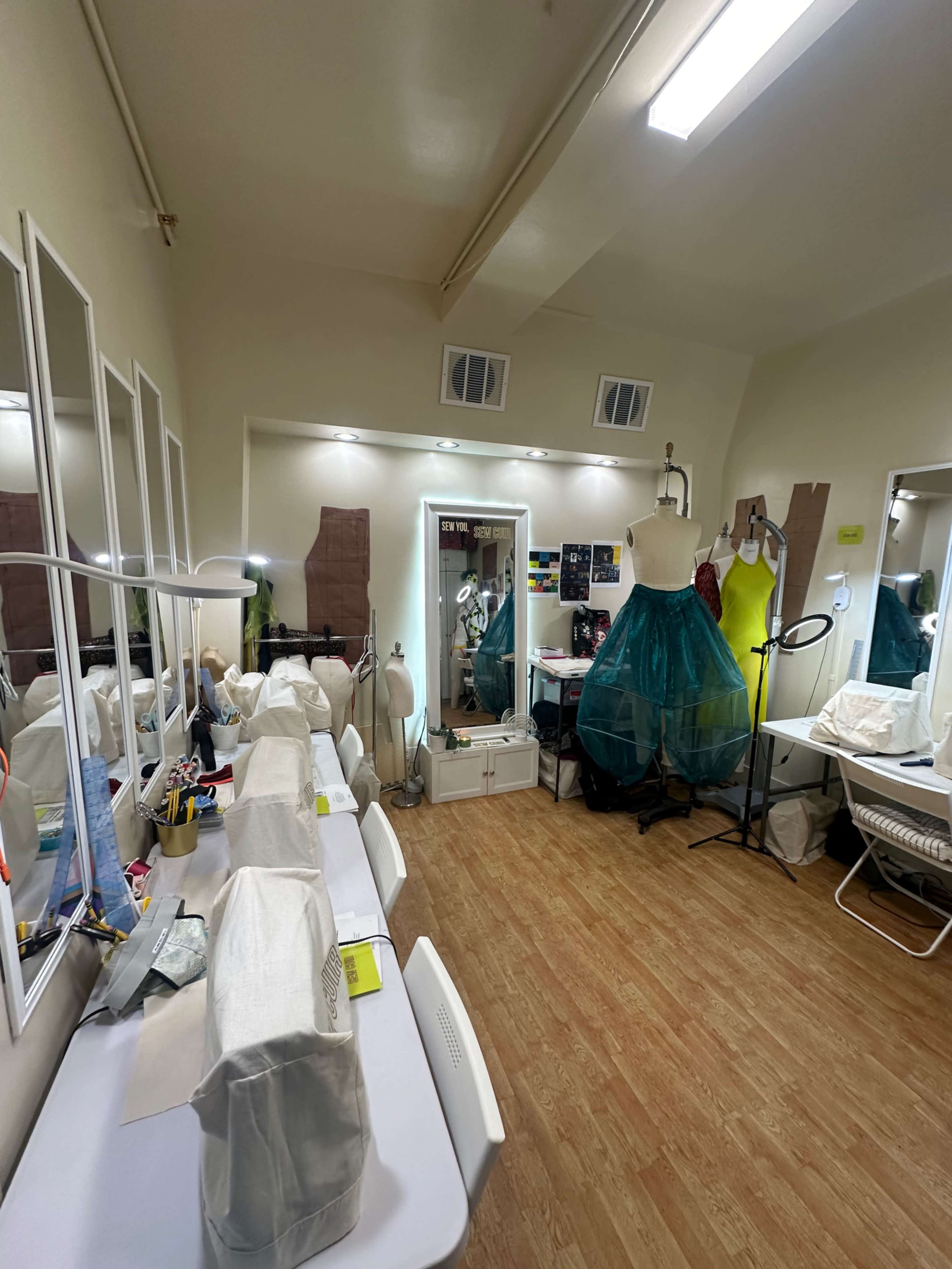 The image shows a brightly lit sewing studio with multiple mirrors, sewing stations, a dress form, and a colorful gown hanging on a rack.