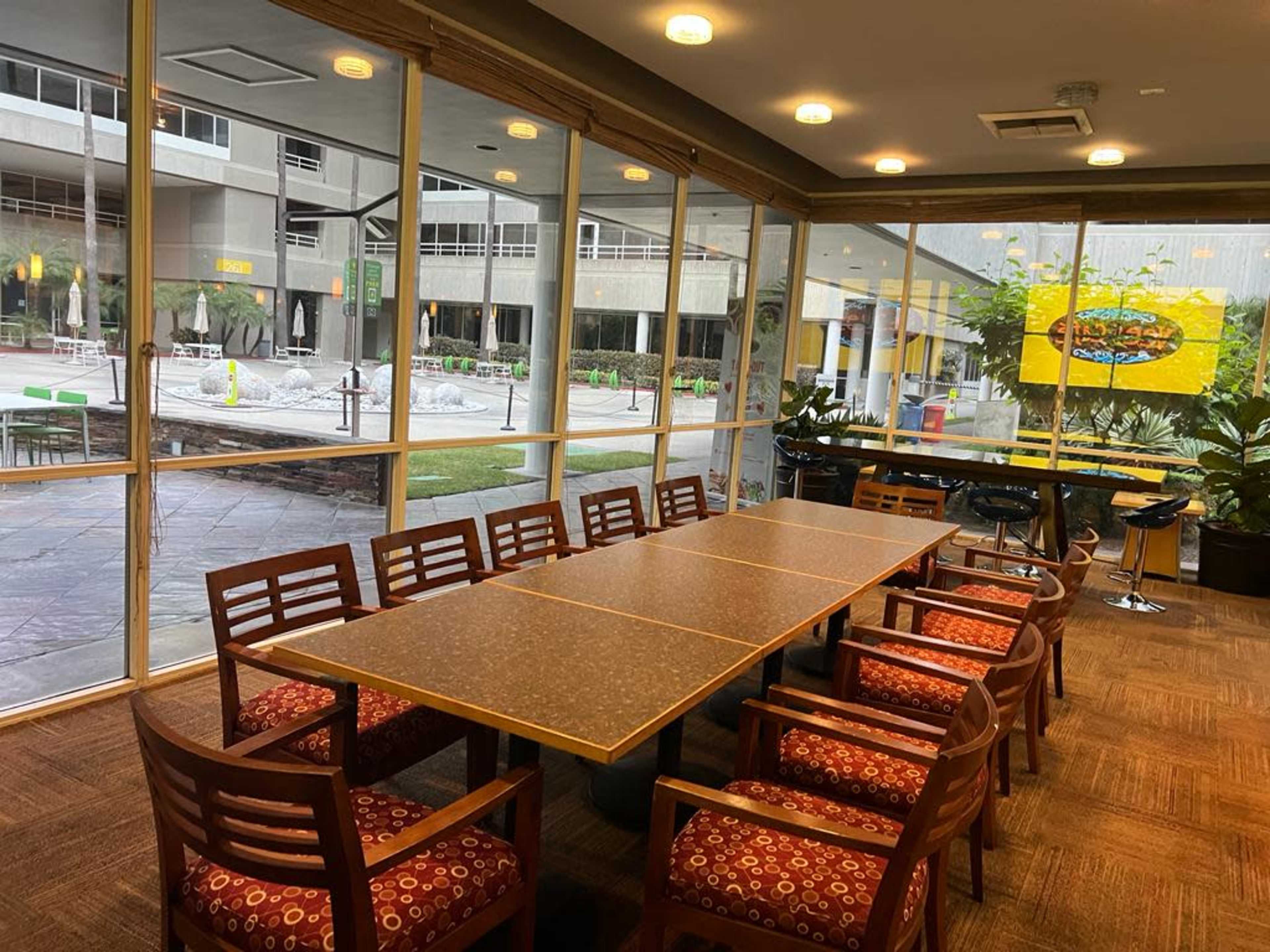 A large conference table with wooden chairs is set up in a glass-enclosed room overlooking an outdoor area with greenery and seating.