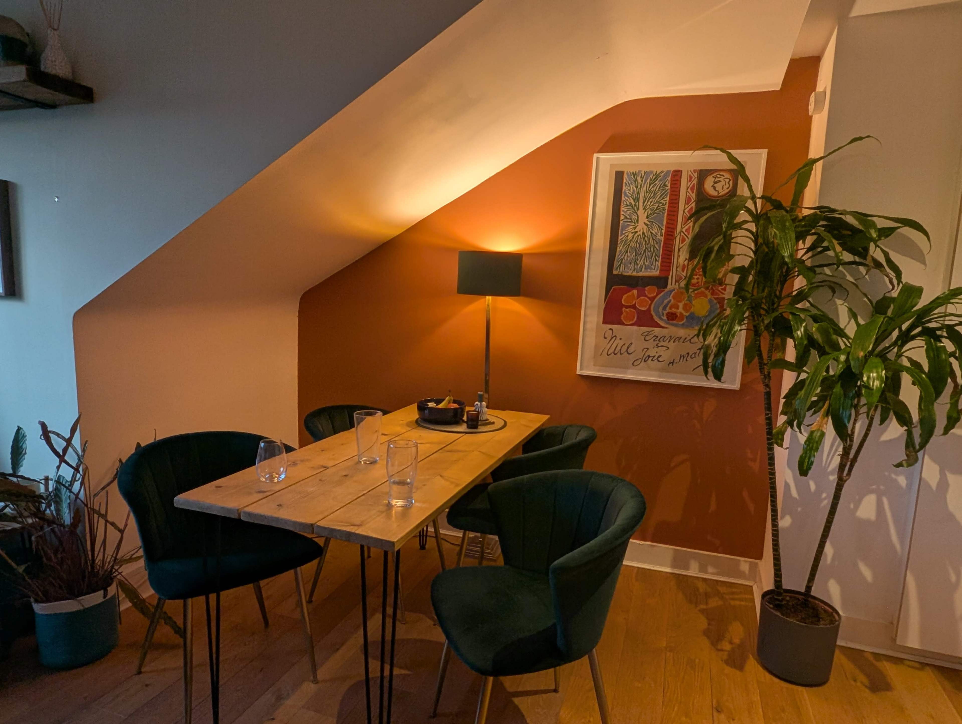 The image shows a cozy dining area with a wooden table, four green chairs, a floor lamp, and a large plant under a sloped ceiling.