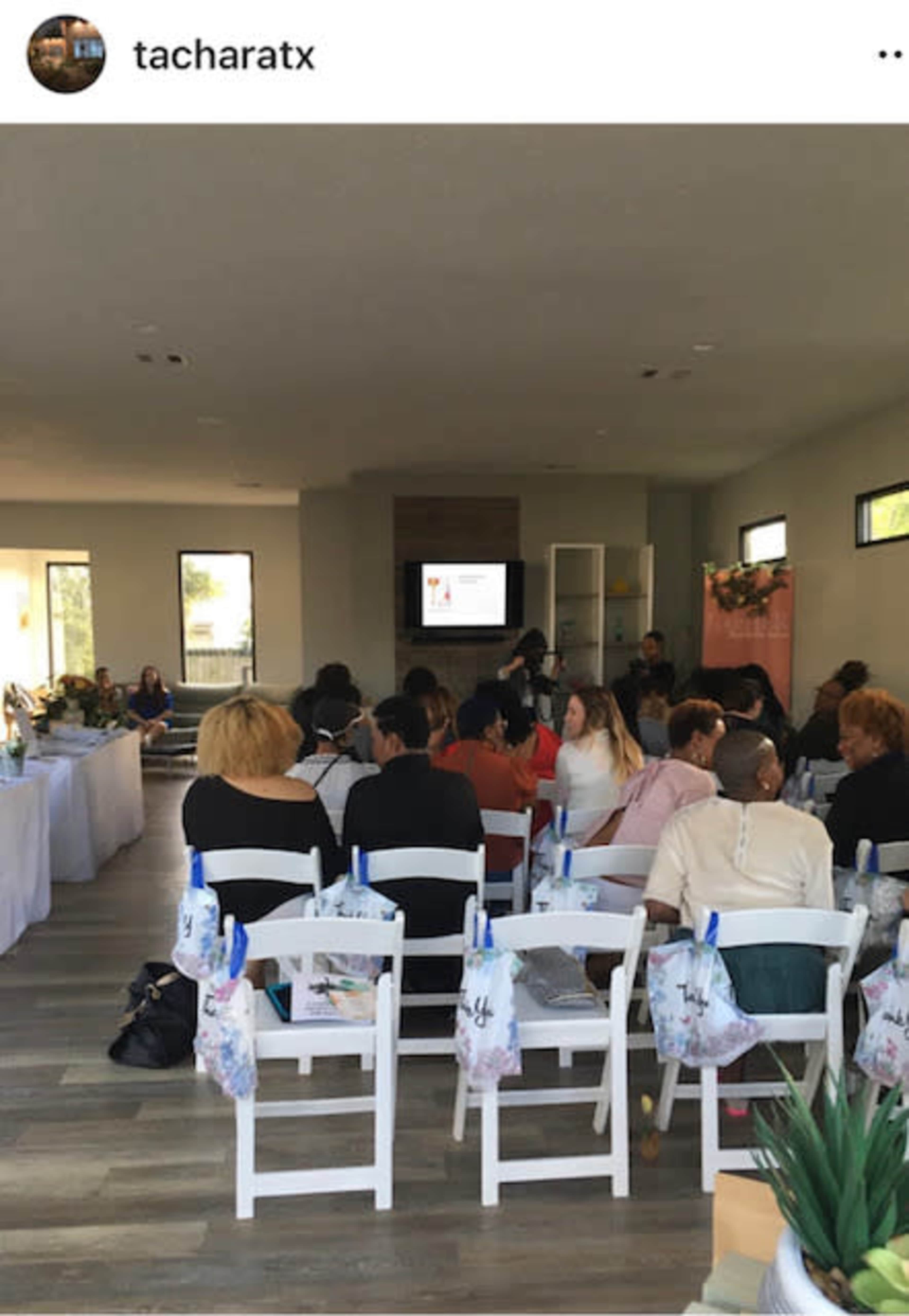 A group of people is seated in a modern room with a large screen at the front, where an event or presentation is taking place.