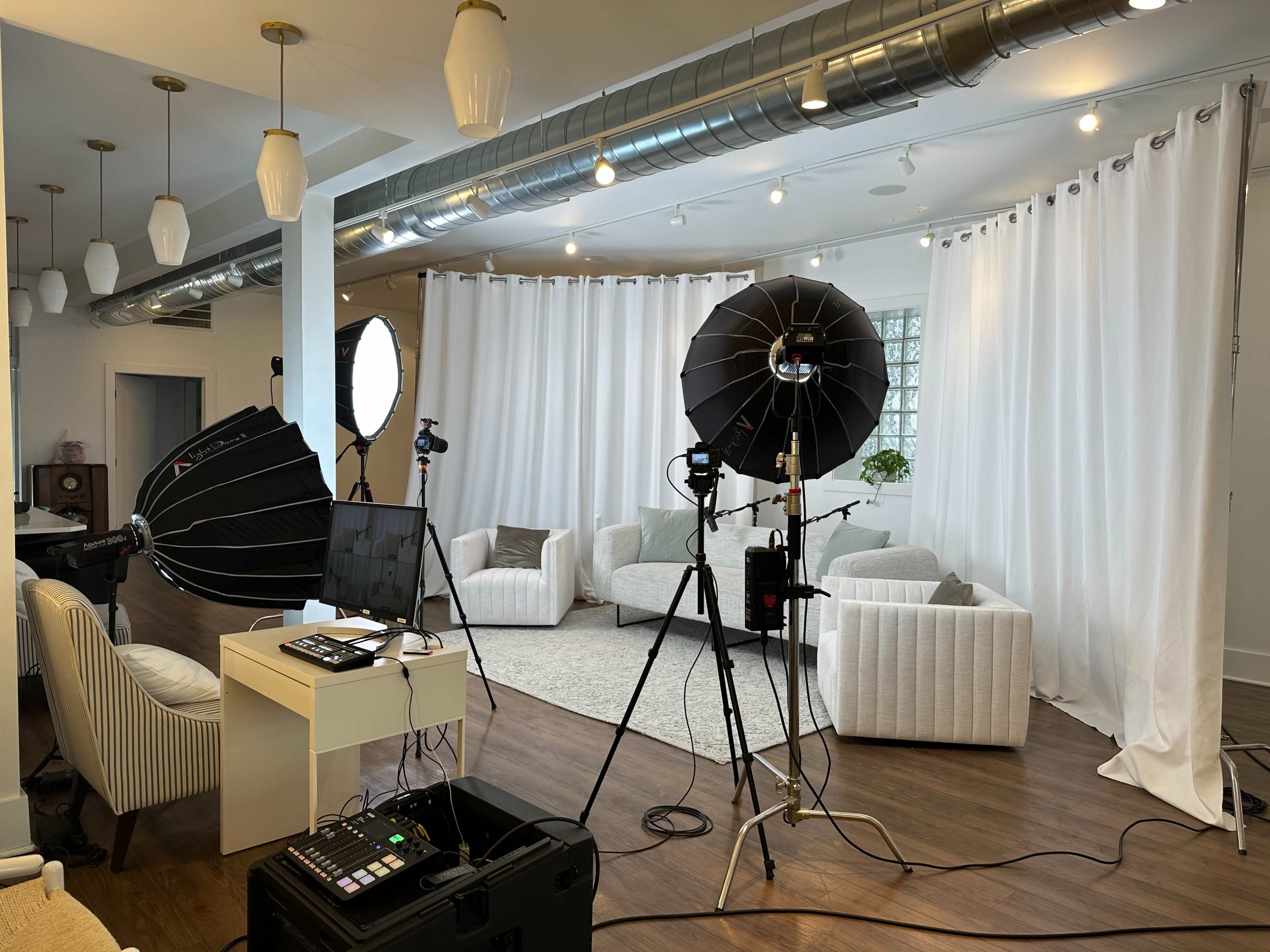 The image shows a photography studio setup with softbox lights, a camera, and a white draped backdrop, arranged around several white upholstered chairs.