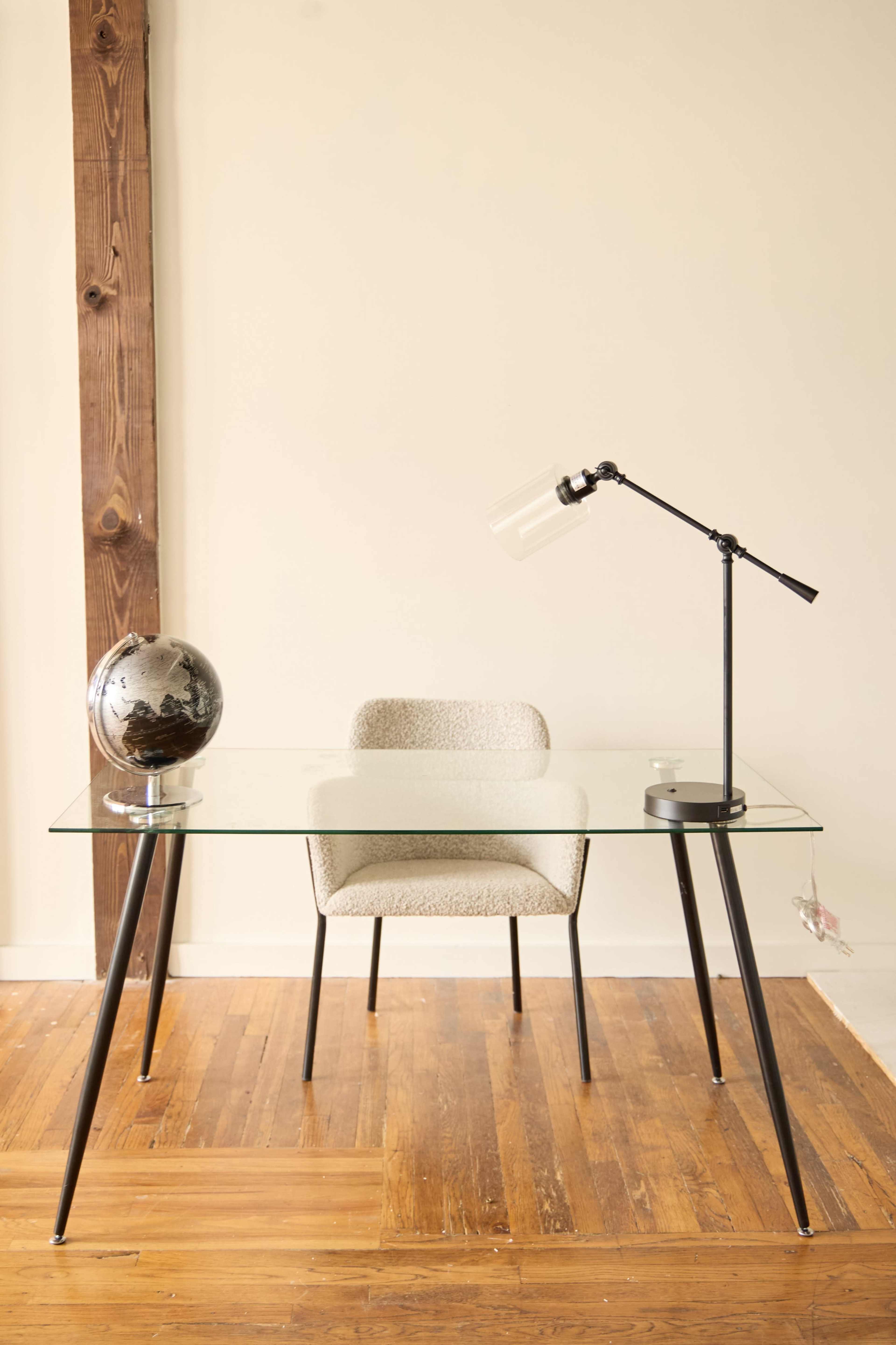 A glass-top desk with a metal lamp and a globe sits next to a chair on a wooden floor in a minimalist room.