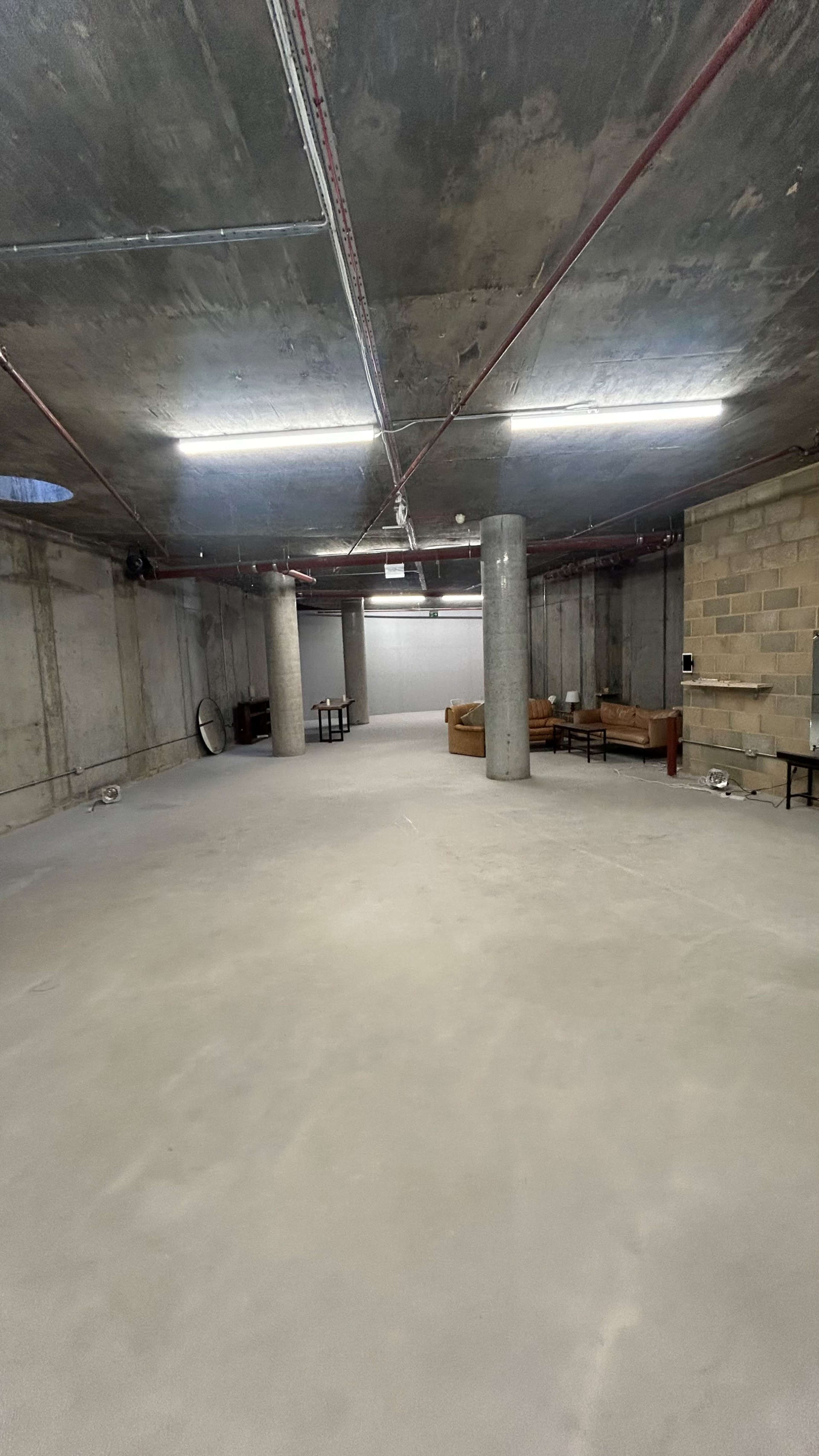 The image shows a spacious underground room with concrete walls, exposed pipes, and minimal furniture, including two couches and several small tables.