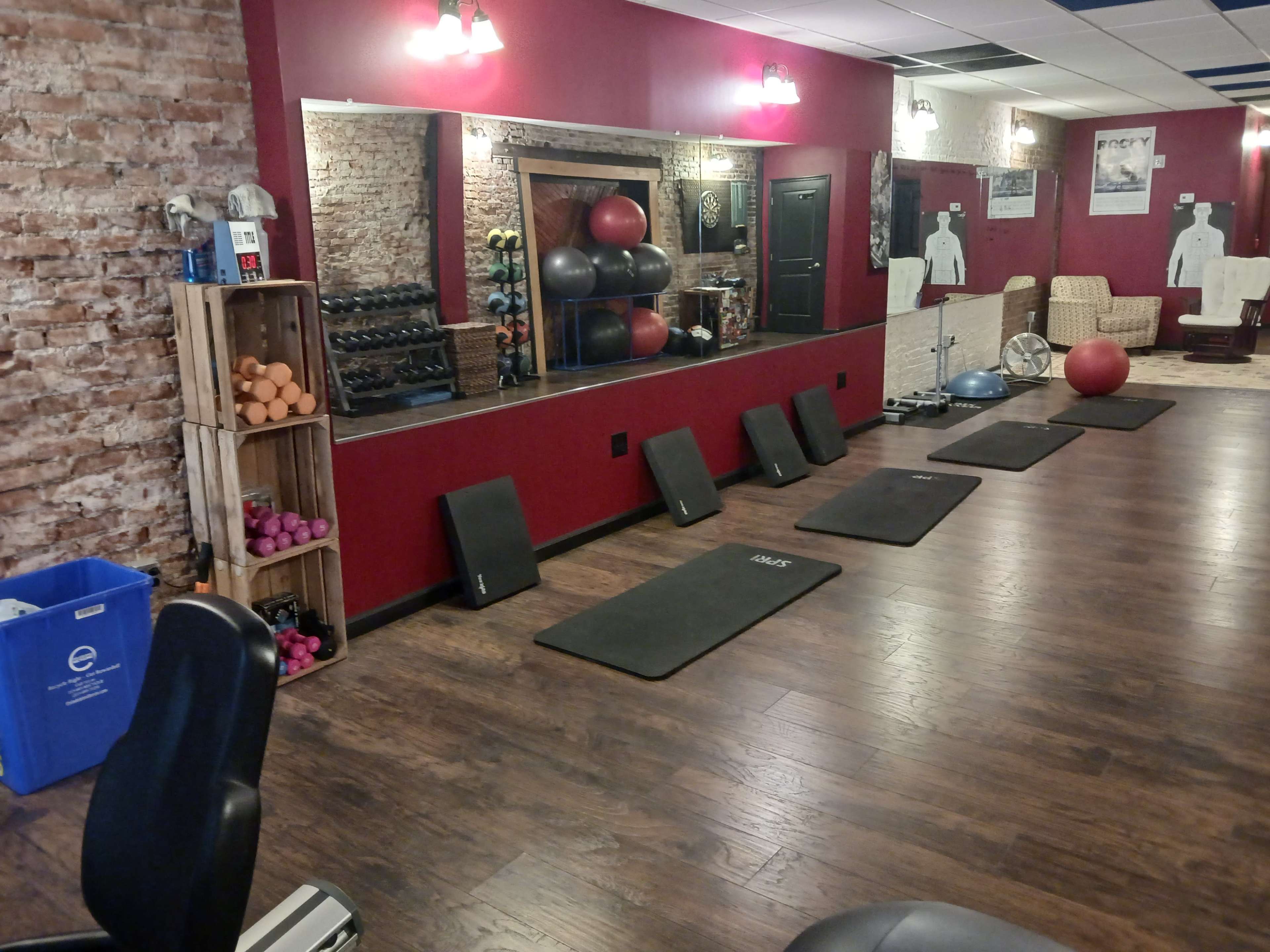 The image shows a fitness studio with exercise mats arranged on the floor, weights on a rack, and a large mirror reflecting the workout area.