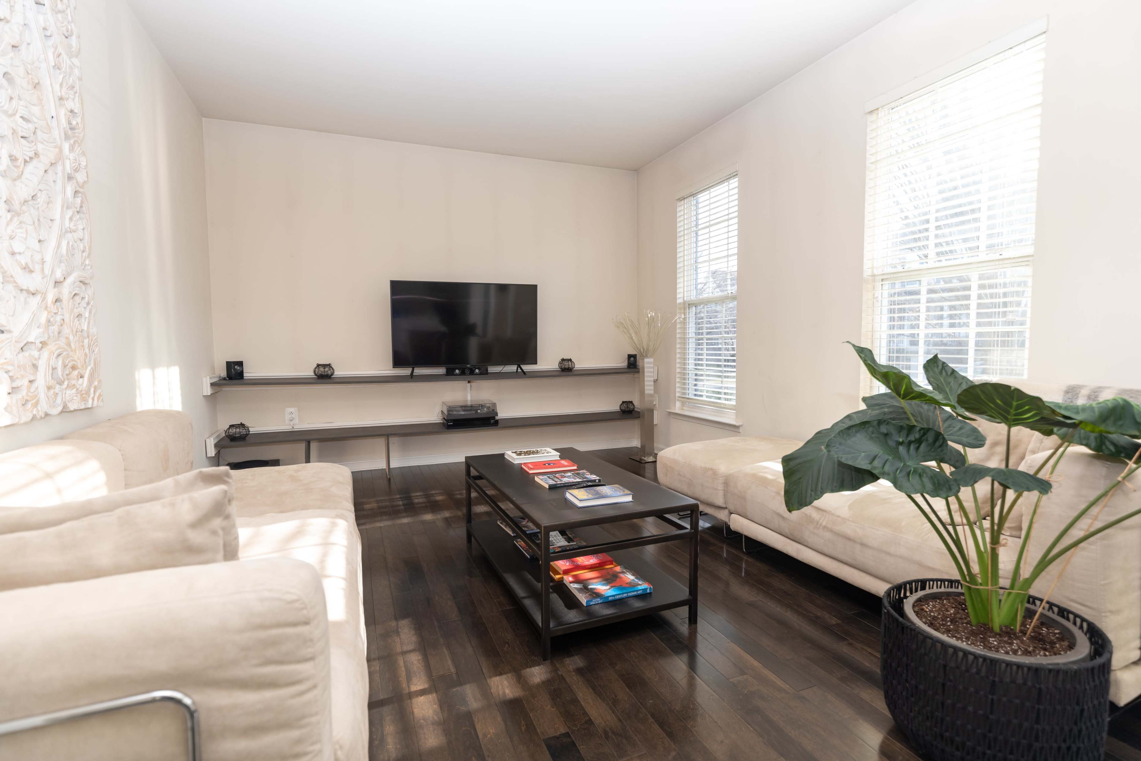 A modern living room features a flat-screen TV mounted on the wall, a low coffee table with magazines, and two beige sofas around it, with a potted plant in the corner.