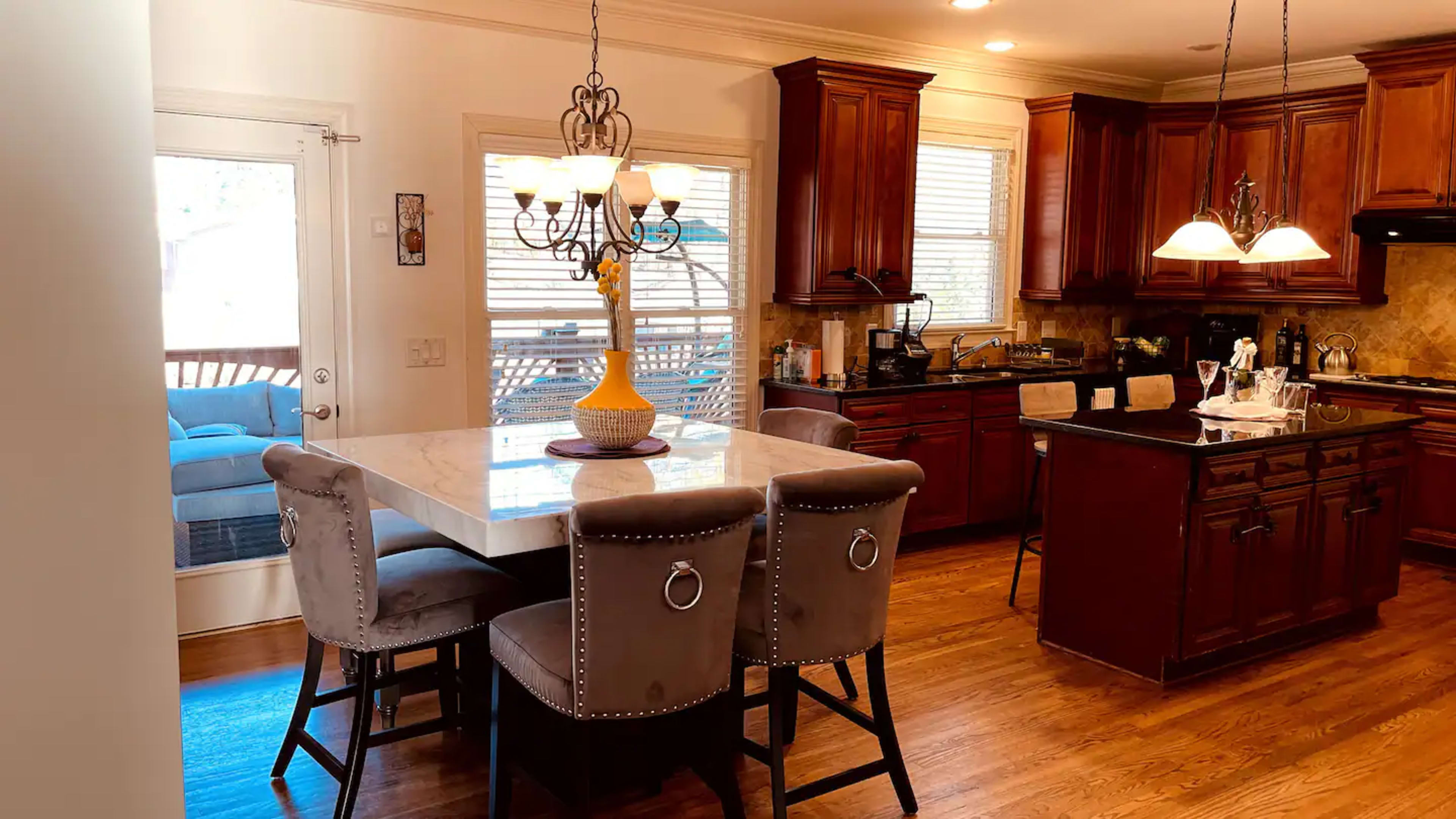 The image shows a modern kitchen with dark wooden cabinets, a large island with seating, and an adjoining dining area illuminated by a chandelier.