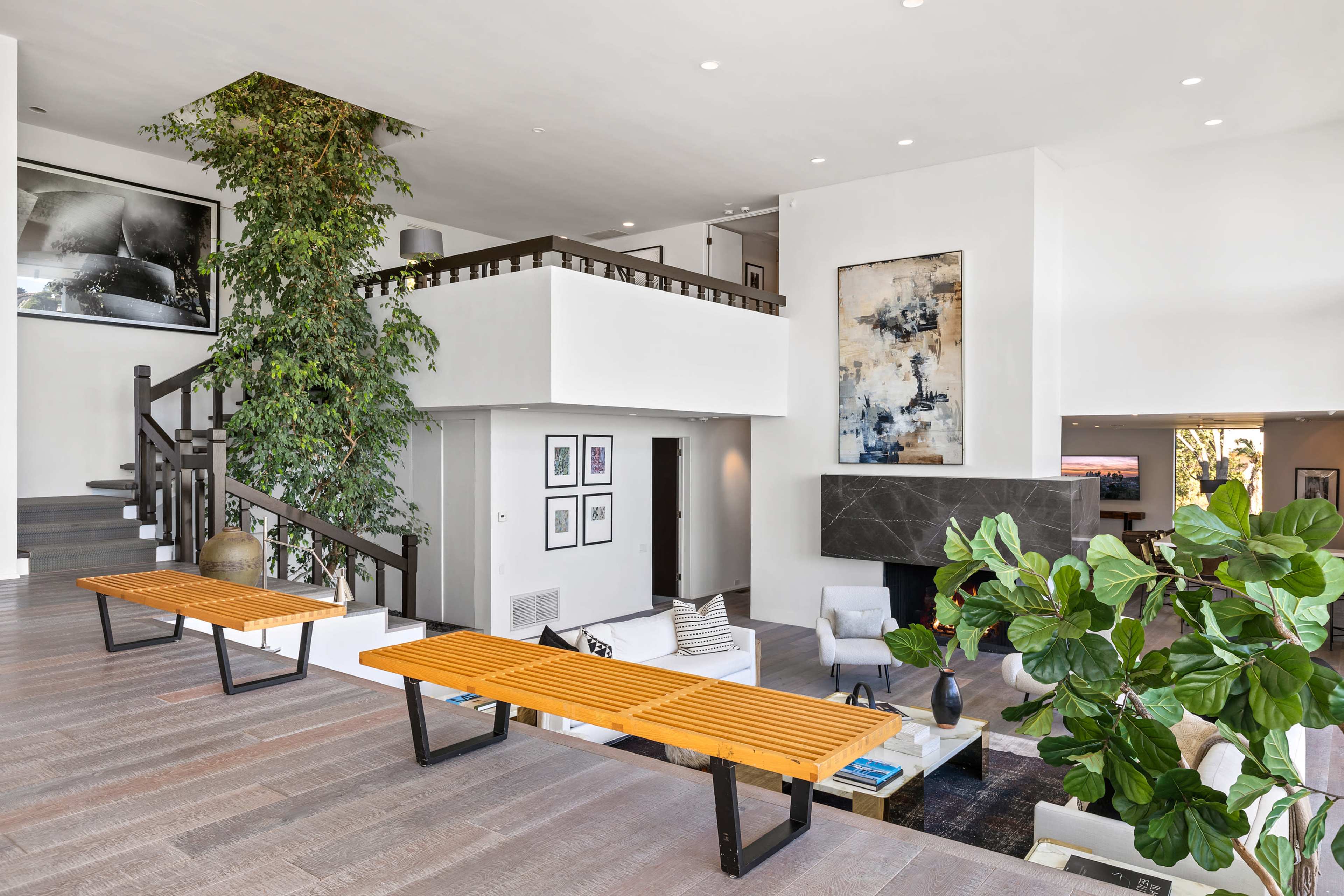 A spacious, modern living area with a two-story design, featuring wooden benches, a large indoor plant, and contemporary art pieces on the walls.