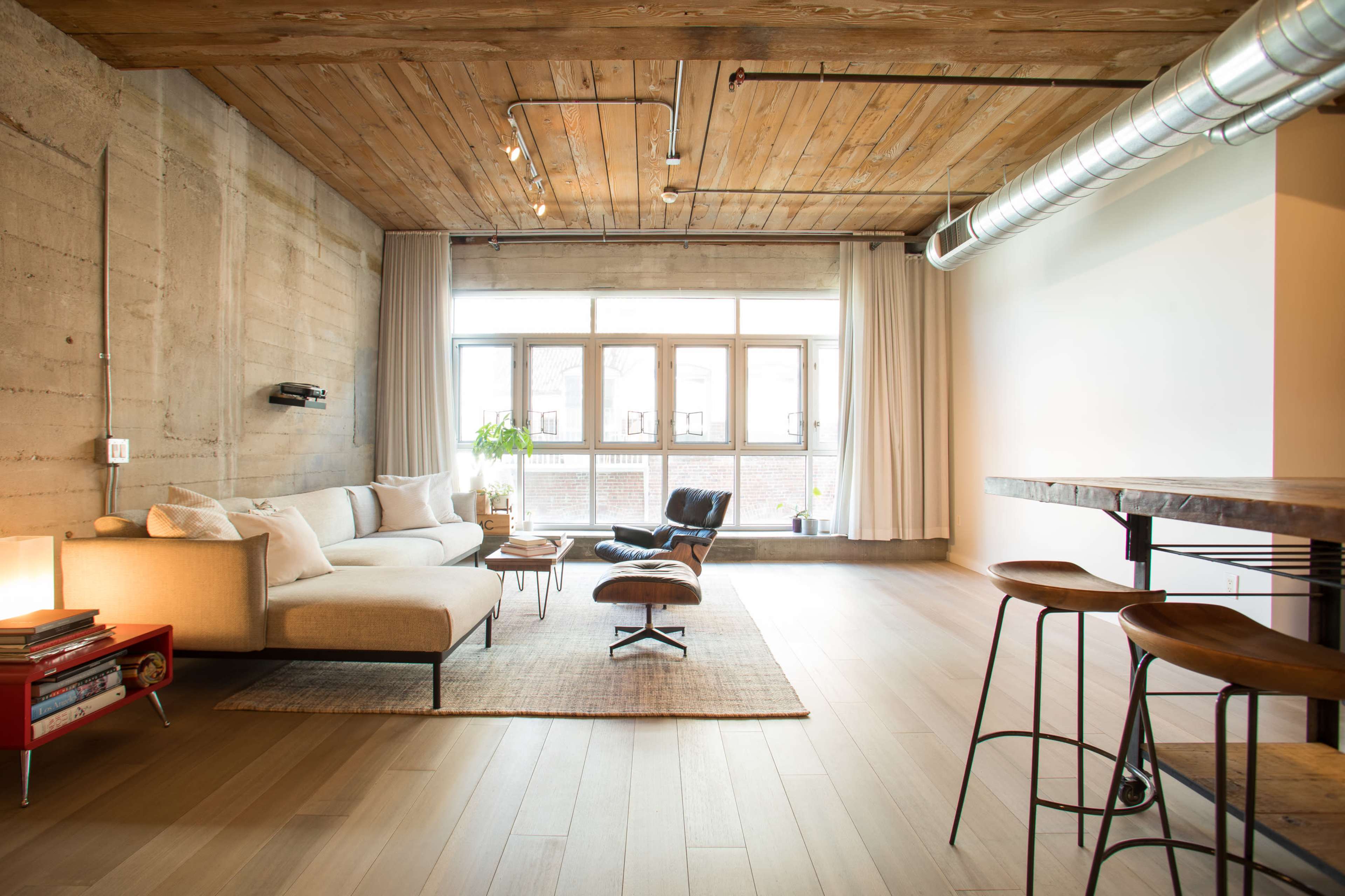 The image shows a modern living room with a concrete wall, wooden ceiling, large windows, a beige sectional sofa, an Eames-style lounge chair, and a dining area with metal bar stools.